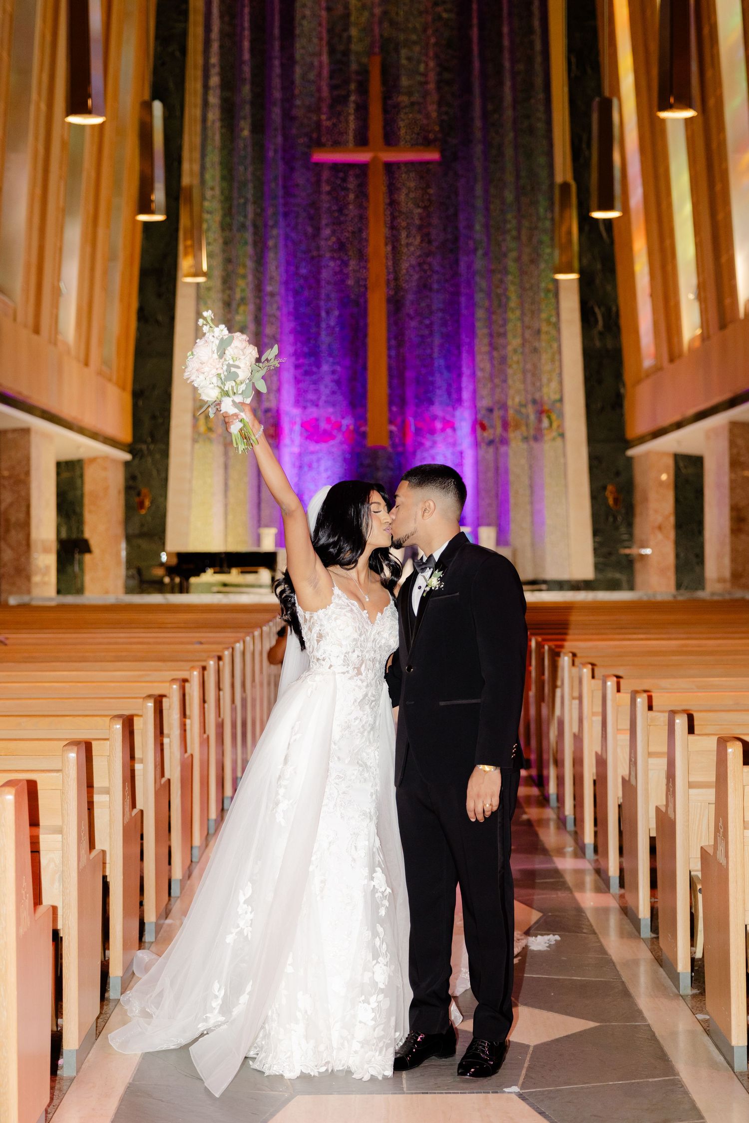 Celebratory wedding moment in church aisle with raised bouquet and purple cross backdrop.