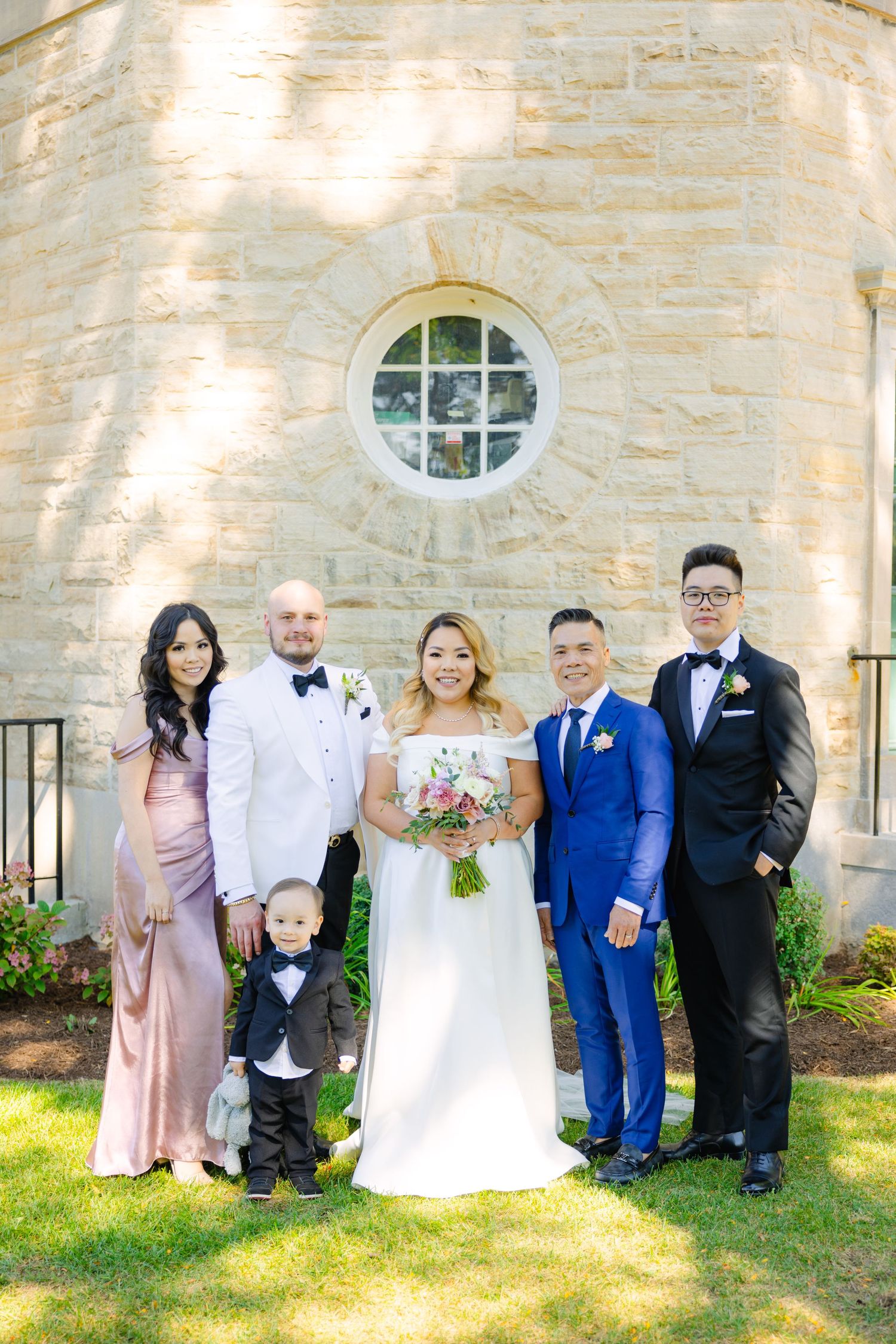 Wedding group with child poses by stone church wall featuring round window and landscaping.