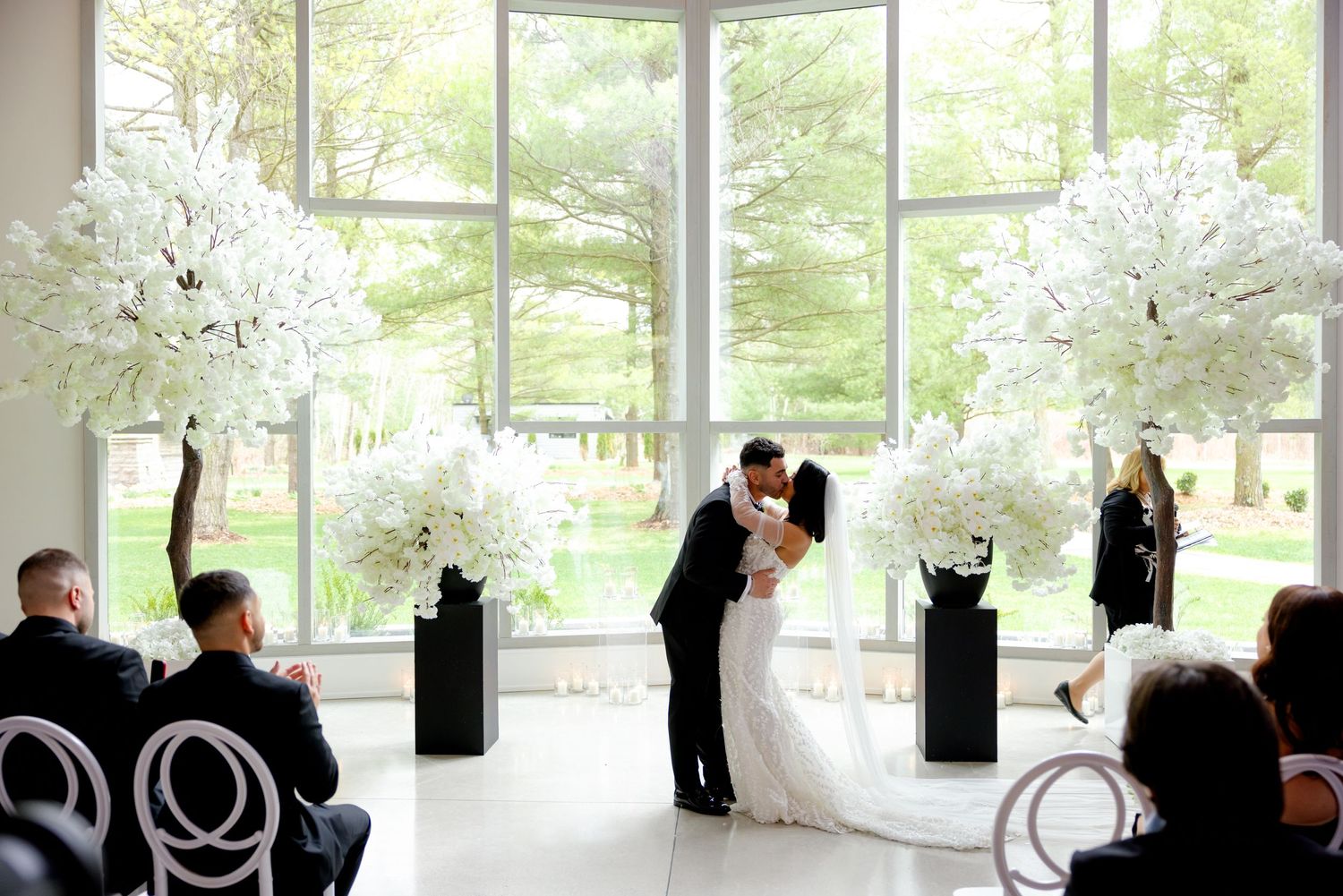 Wedding ceremony in bright venue with guests seated on modern chairs and white floral decor.