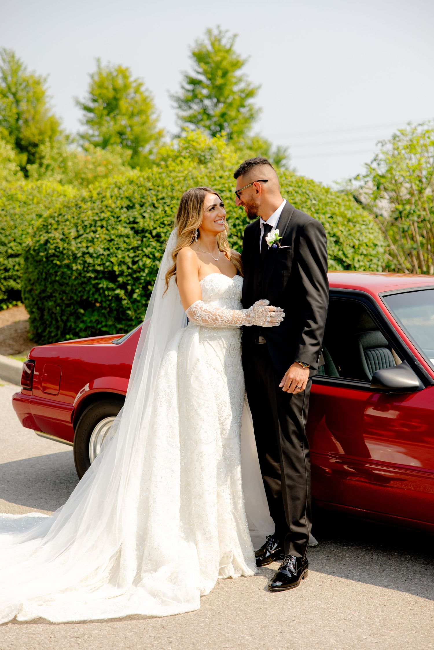 Elegant bride and groom share intimate moment beside red car with flowing veil and manicured grounds.