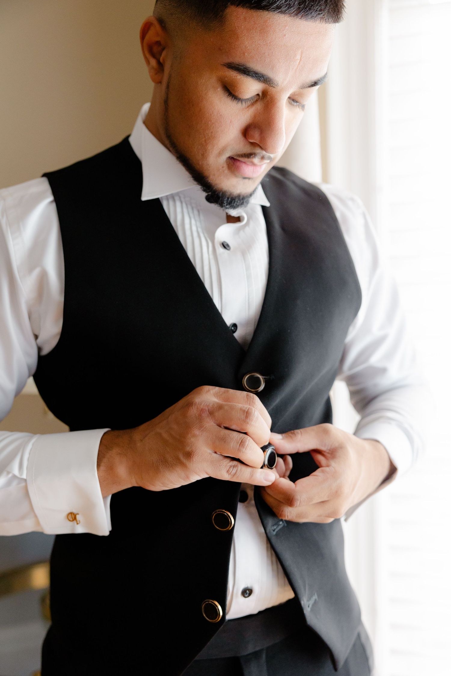 A person buttons up a black formal vest while wearing a pleated white dress shirt.