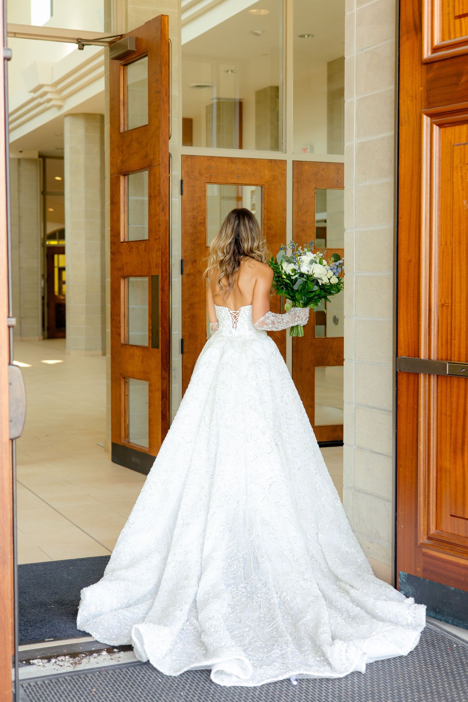 A bride in a ballgown style wedding dress enters through wooden doors carrying a white bouquet.