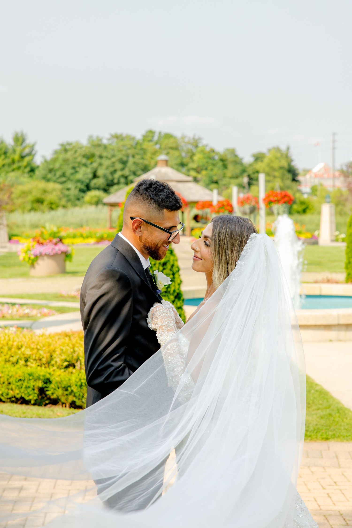 Wedding couple embraces in sunlit garden while long veil flows dramatically in the breeze.