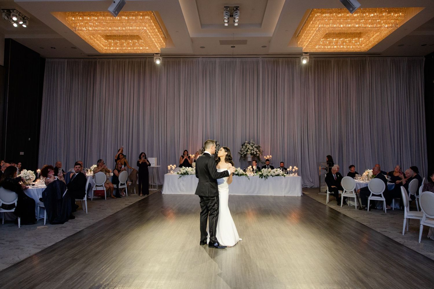 Wide shot of couple dancing in reception hall with guests seated at tables.