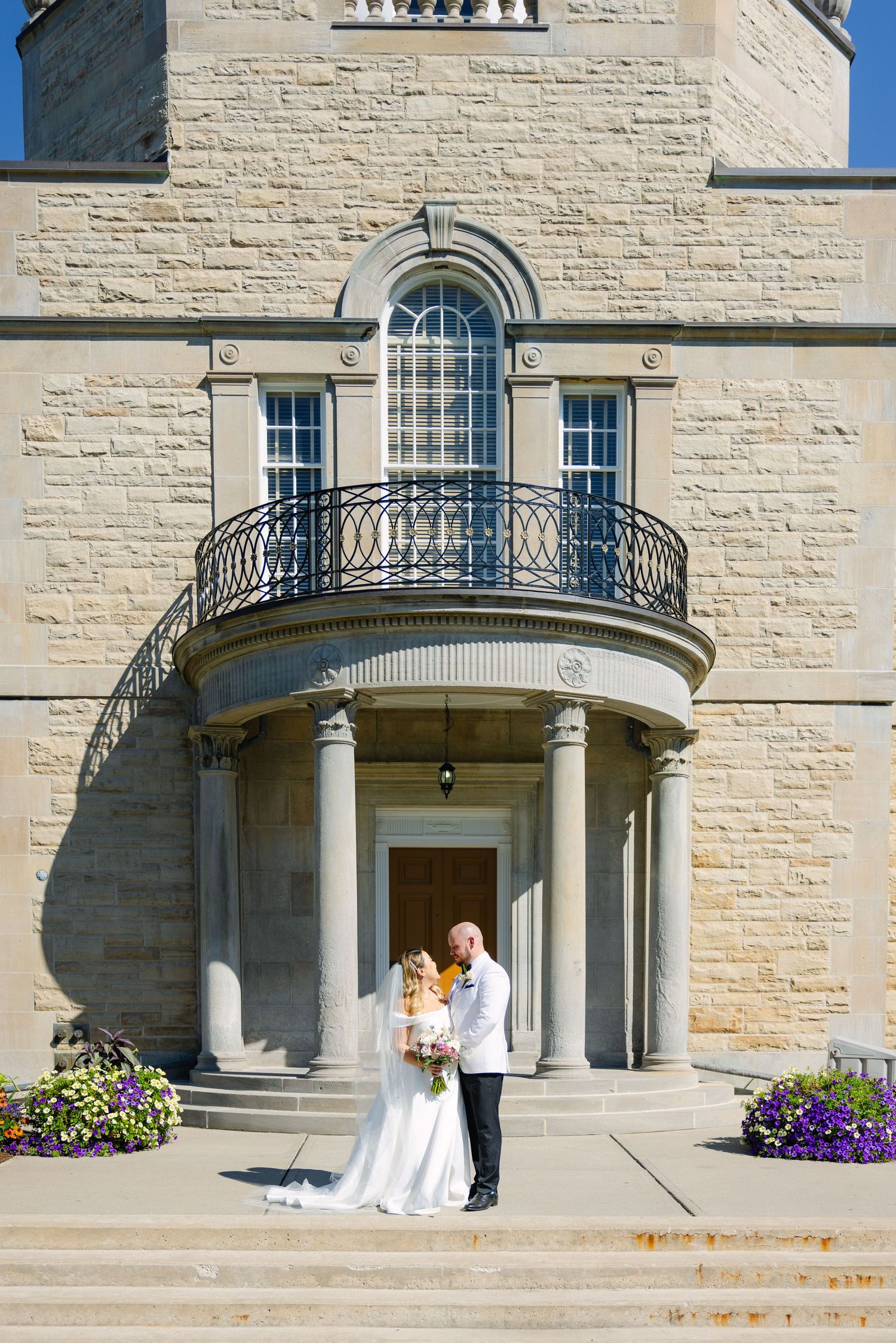 A couple stands at the entrance of a grand limestone building with columns and curved balcony.