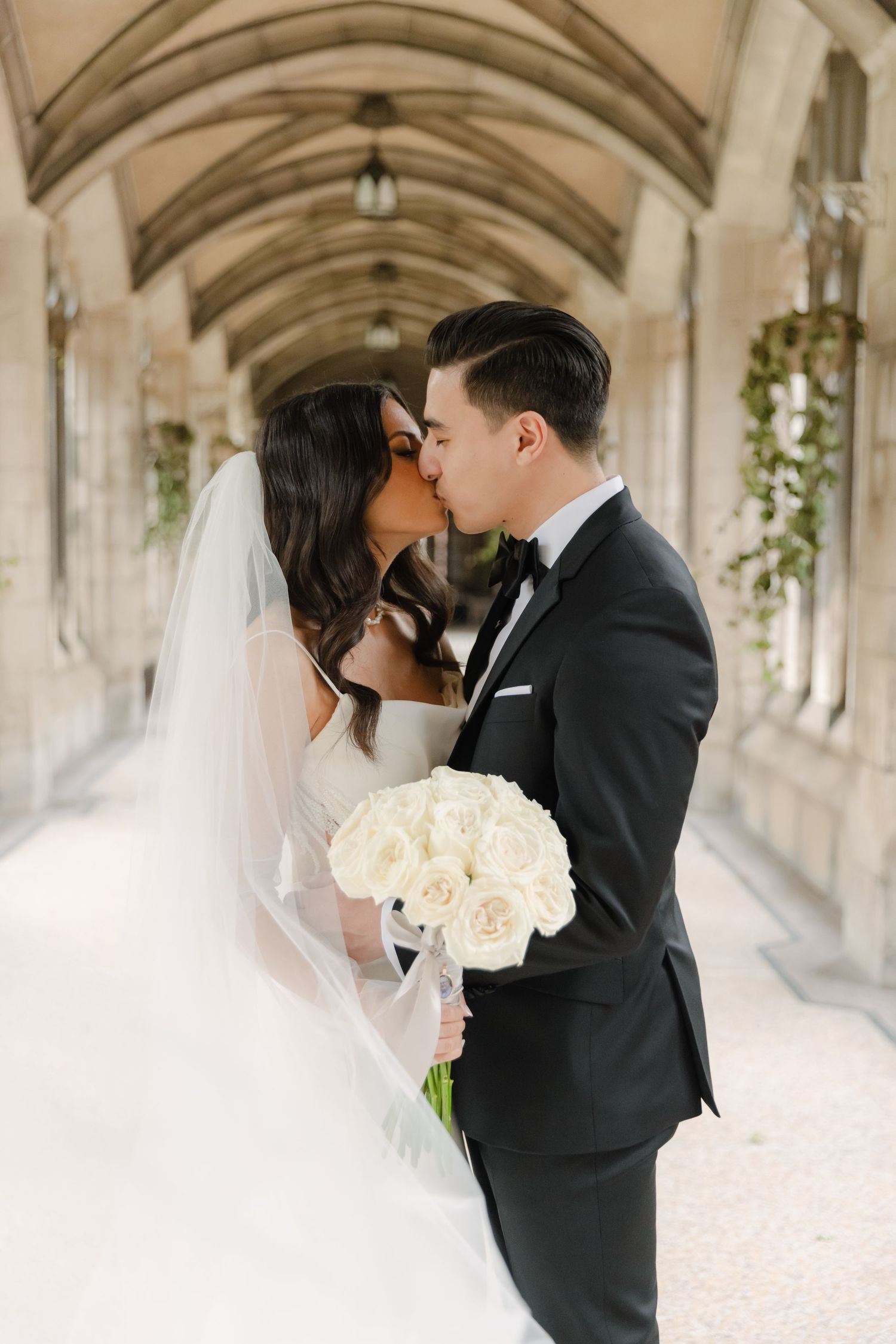 Romantic wedding kiss captured in historic stone hallway with hanging floral decorations.
