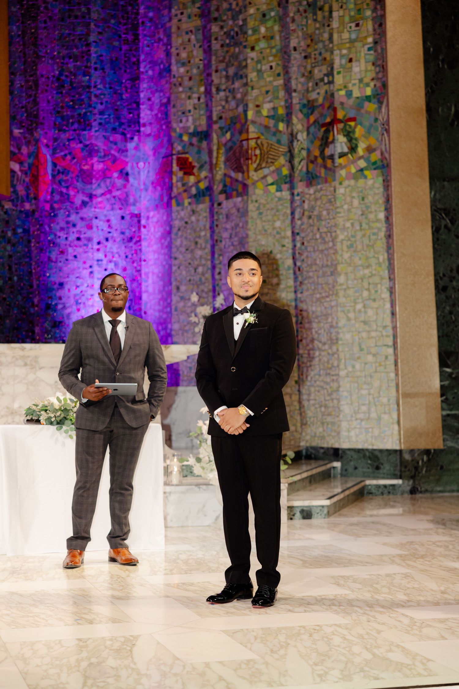 Two men in formal attire stand at church altar with purple lighting backdrop.