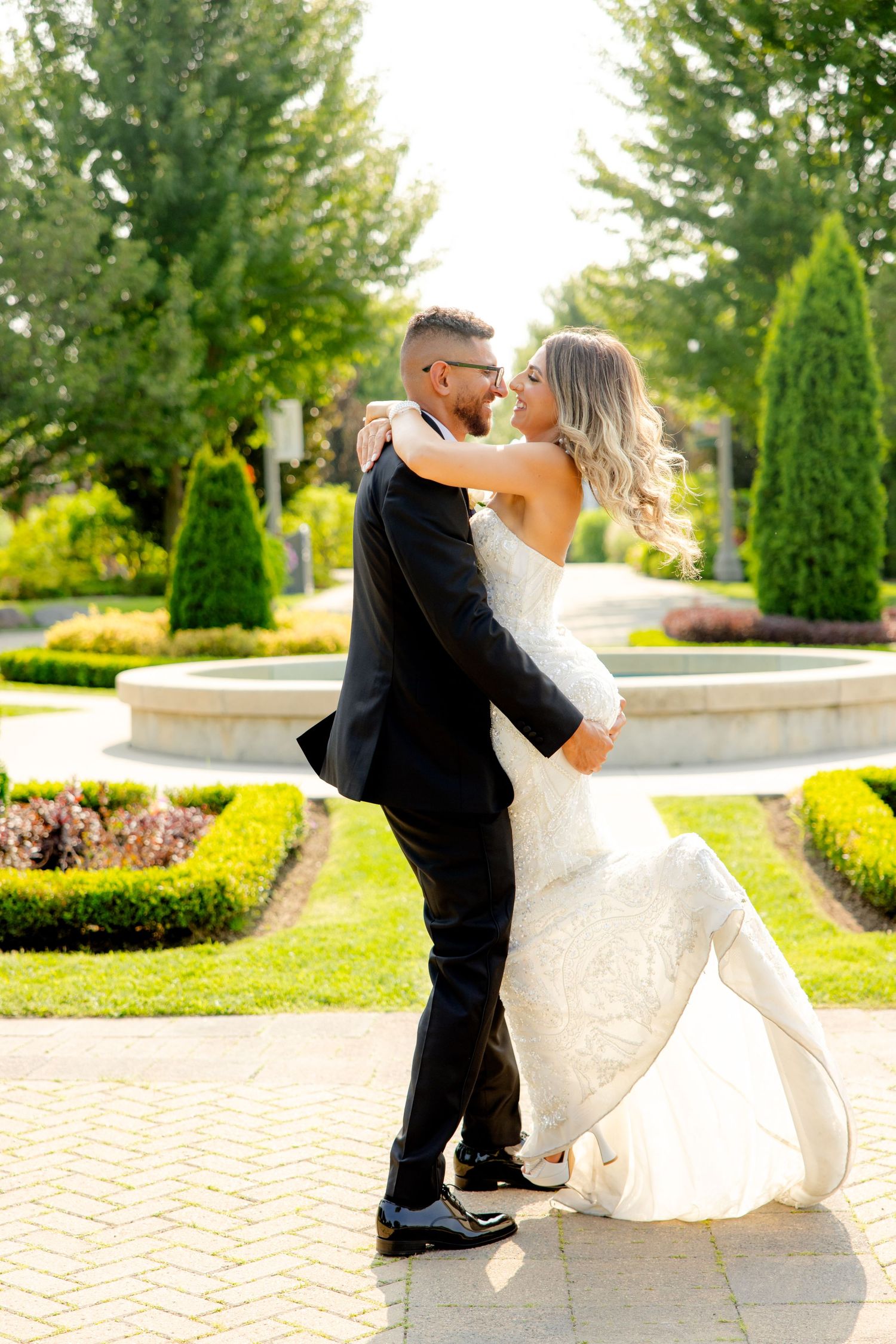 Playful lift pose between bride and groom on brick patio with formal garden landscaping.