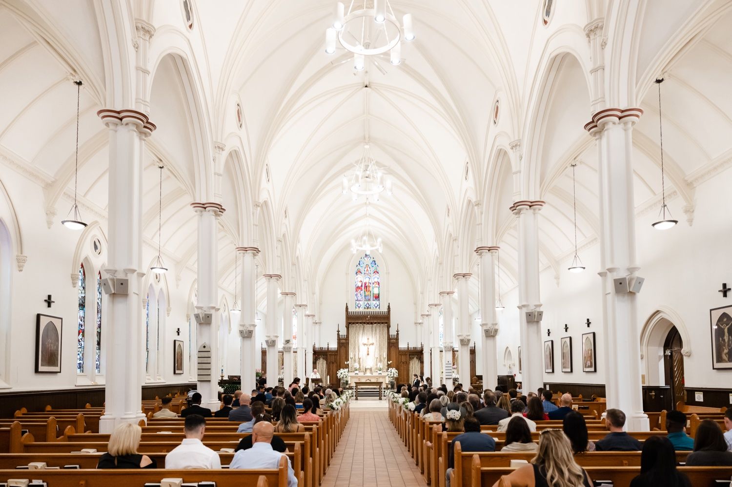 Interior of a grand cathedral with white vaulted ceilings and rows of wooden pews leading to an ornate altar.