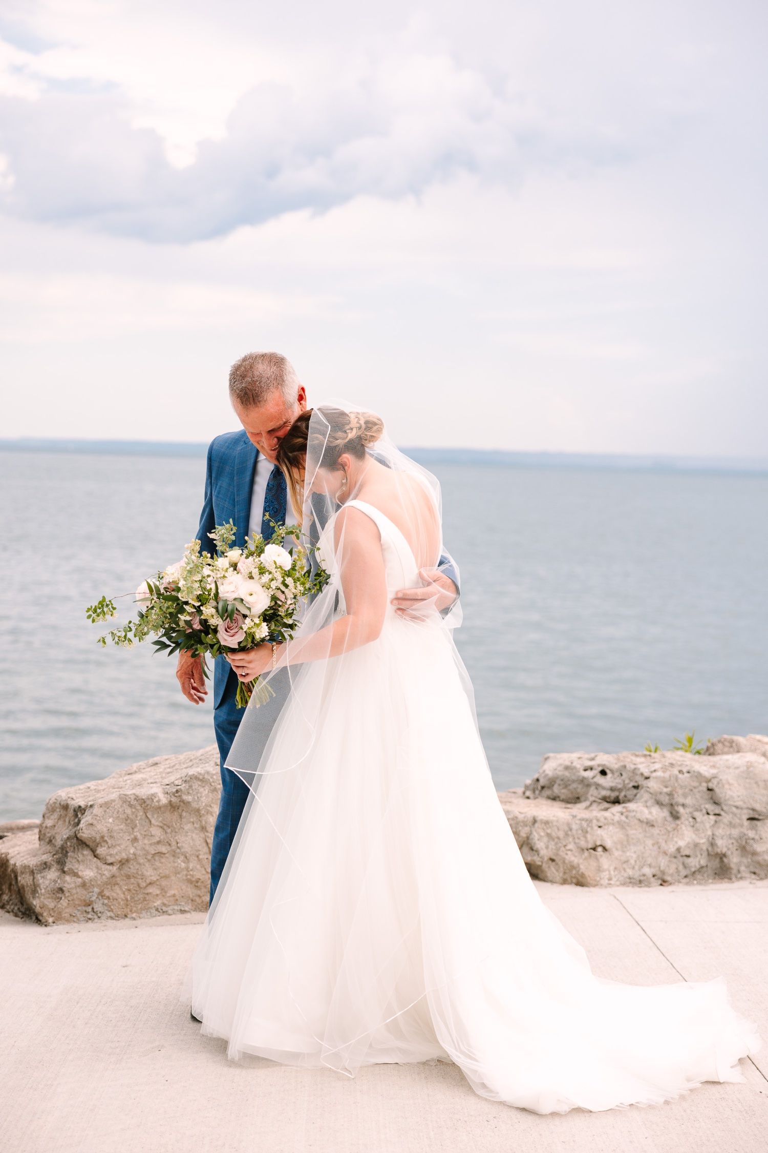 Newlyweds share an intimate moment by the water while holding a lush white and green bouquet.