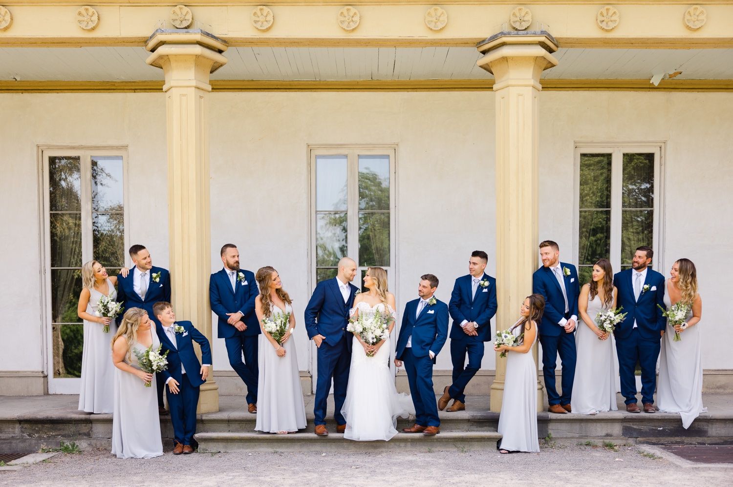 Wedding party arranged casually in front of elegant white colonial style architecture.