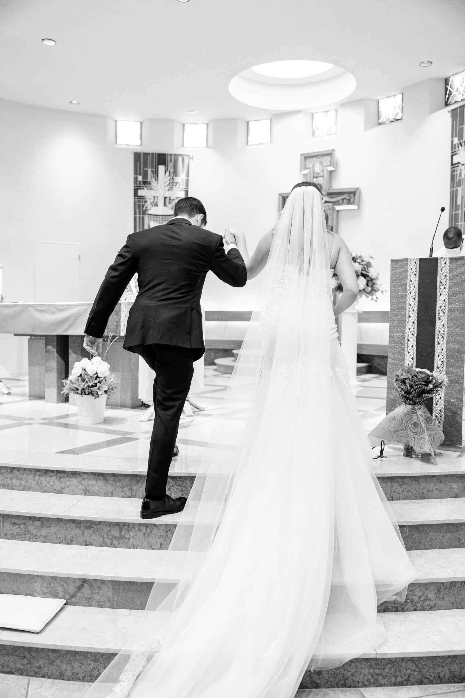 A black and white photo of a couple ascending church altar steps with flowing wedding veil.