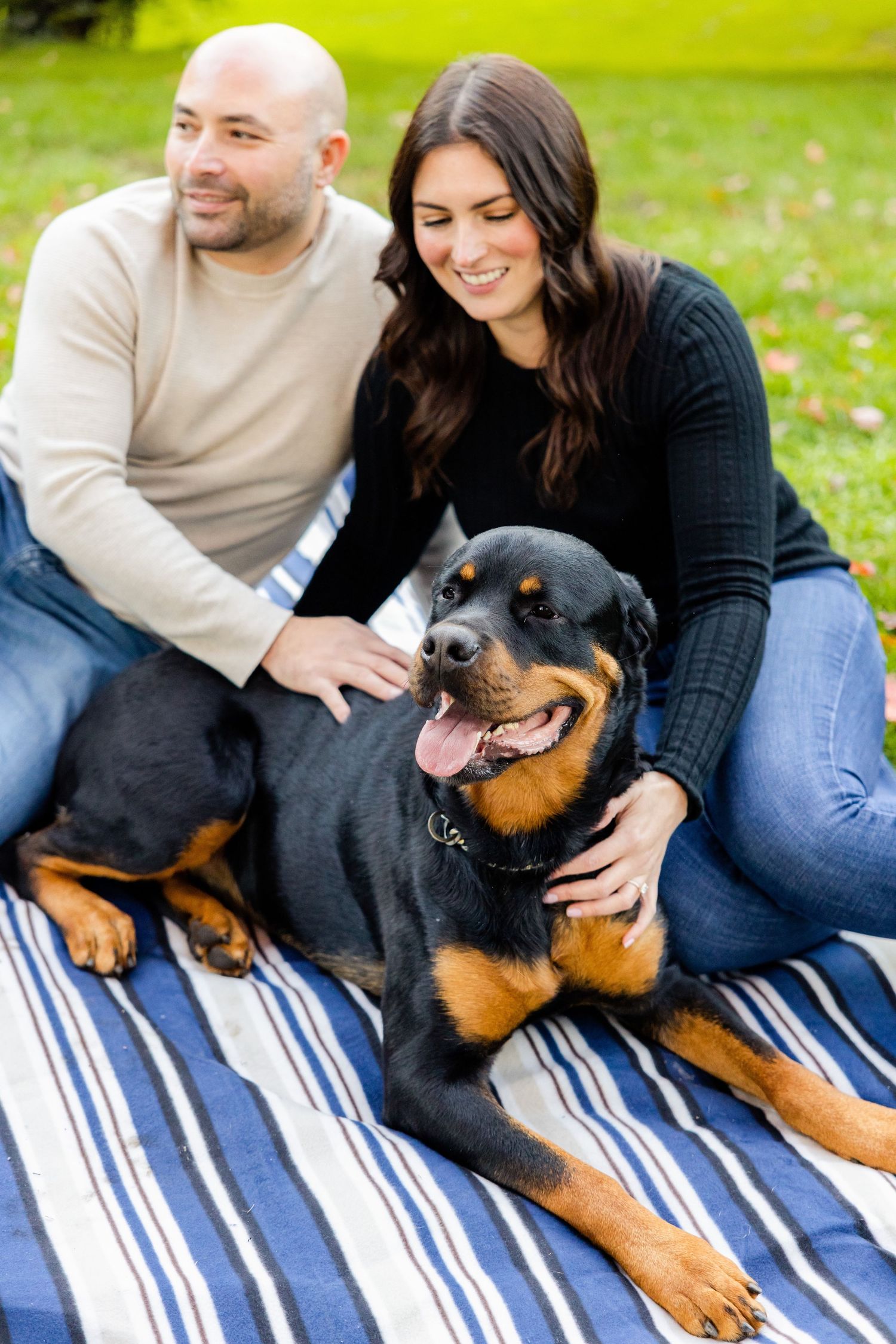 Couple cuddling with smiling Rottweiler dog on blue and white striped blanket outdoors.