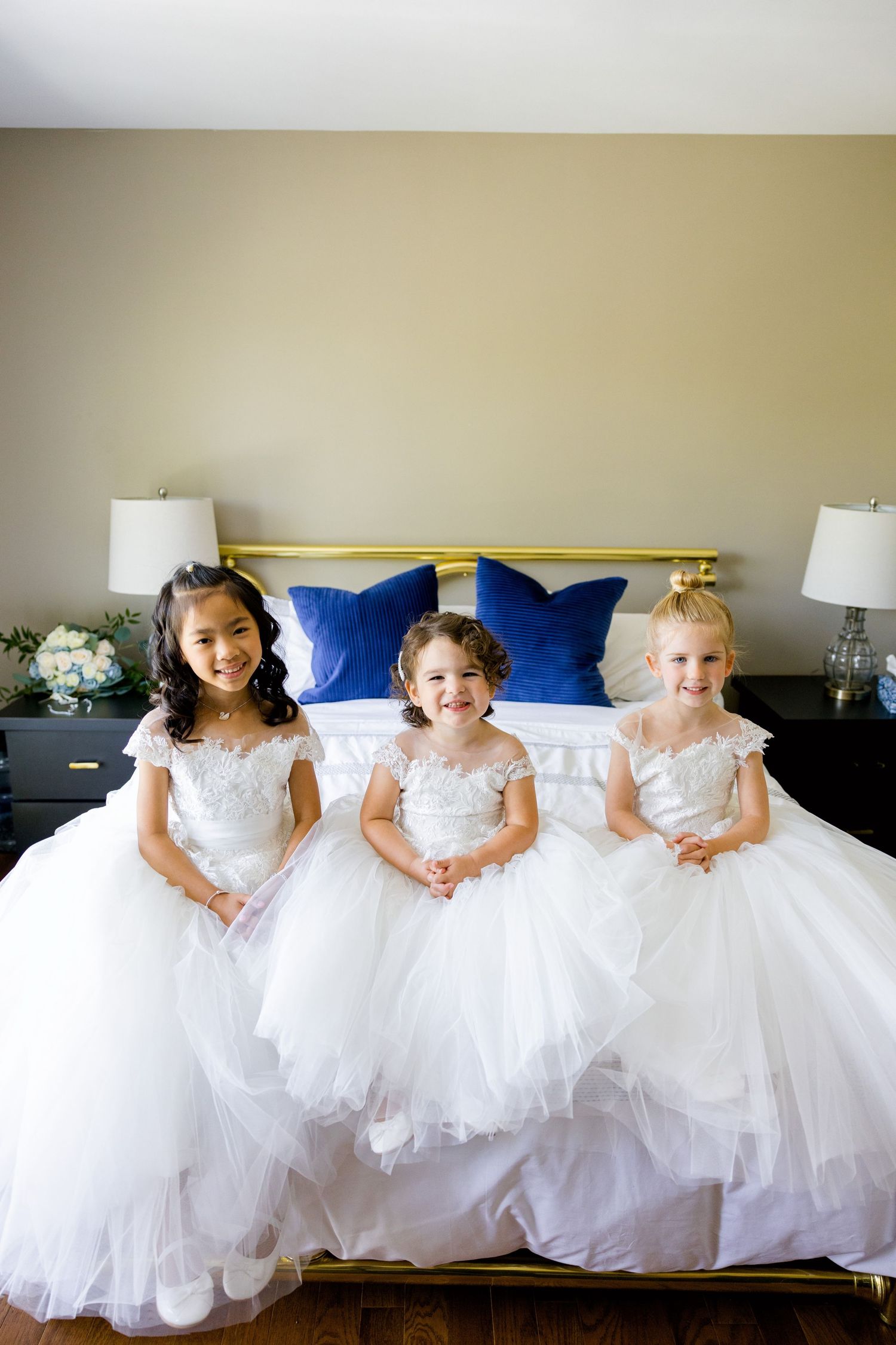 Three flower girls wearing white tulle dresses sitting on a bed with blue pillows.