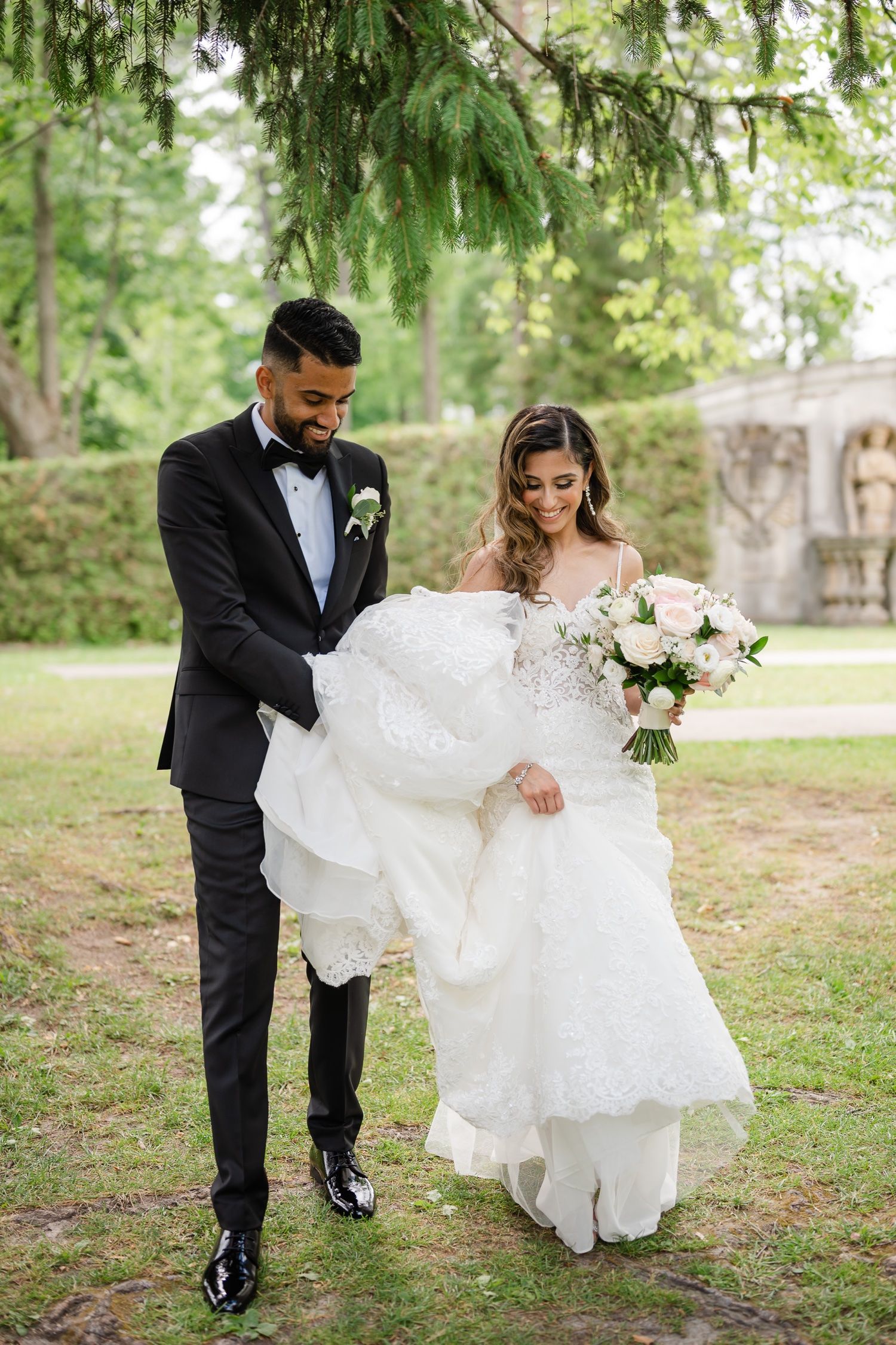 A newlywed couple shares a moment on garden grounds as the groom helps with the bride's dress.