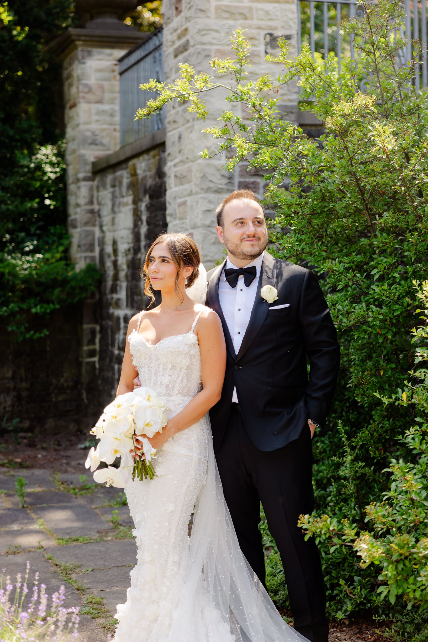 A romantic outdoor wedding portrait taken in front of a rustic stone wall surrounded by blooming flowers.