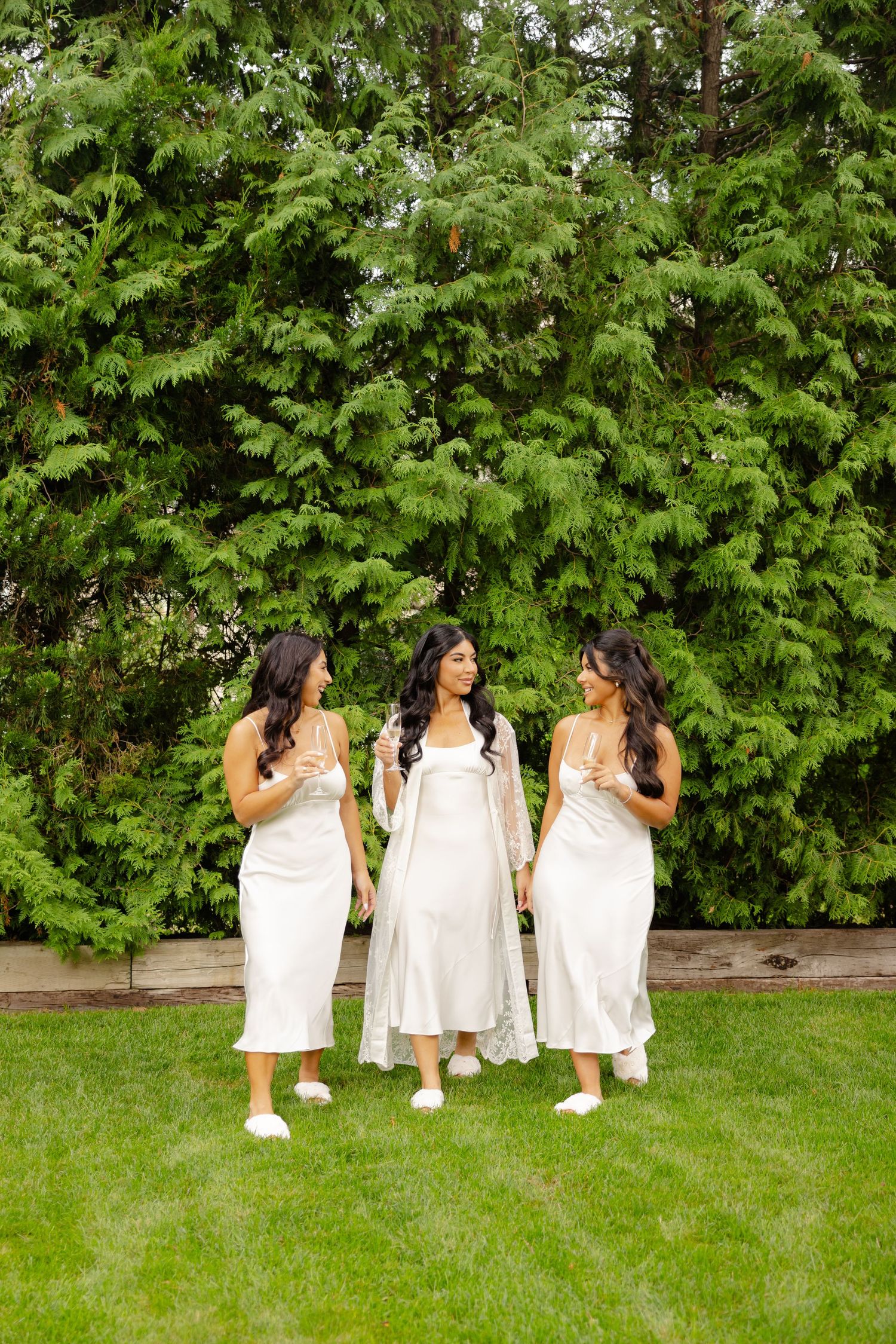 Friends in matching white outfits pose playfully on grass with trees in the background.