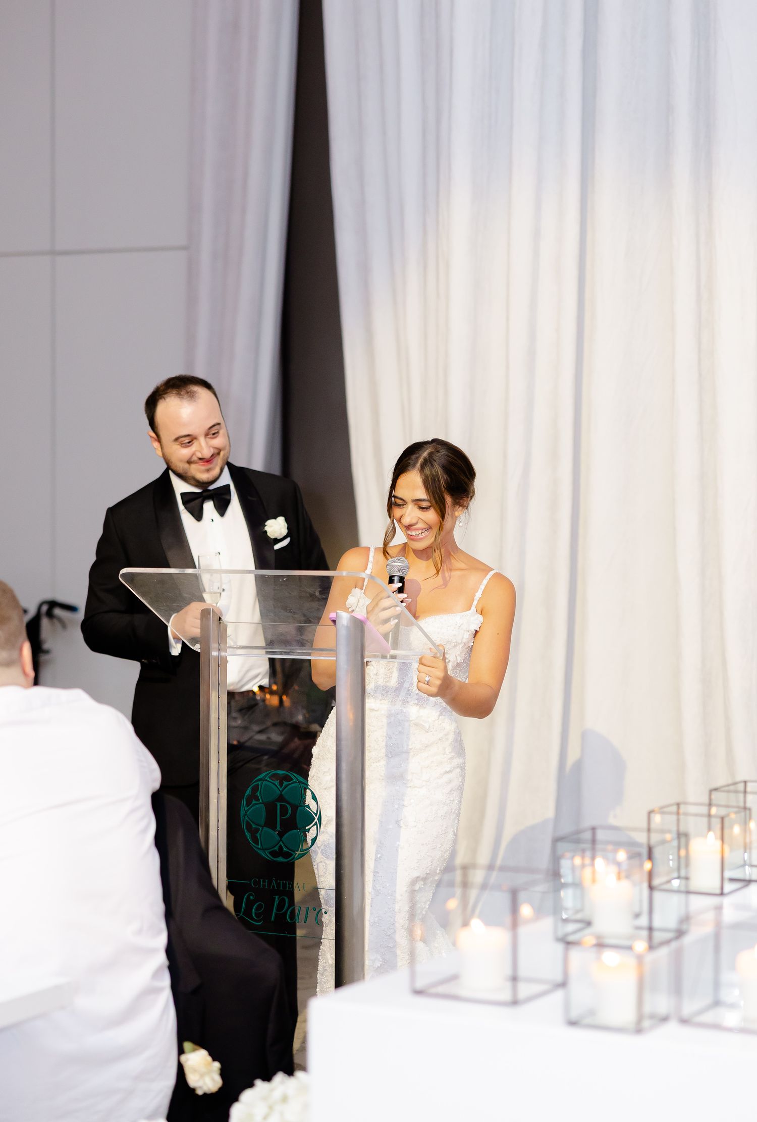 A couple giving speeches at an elegant wedding reception with a clear podium and white curtain backdrop.