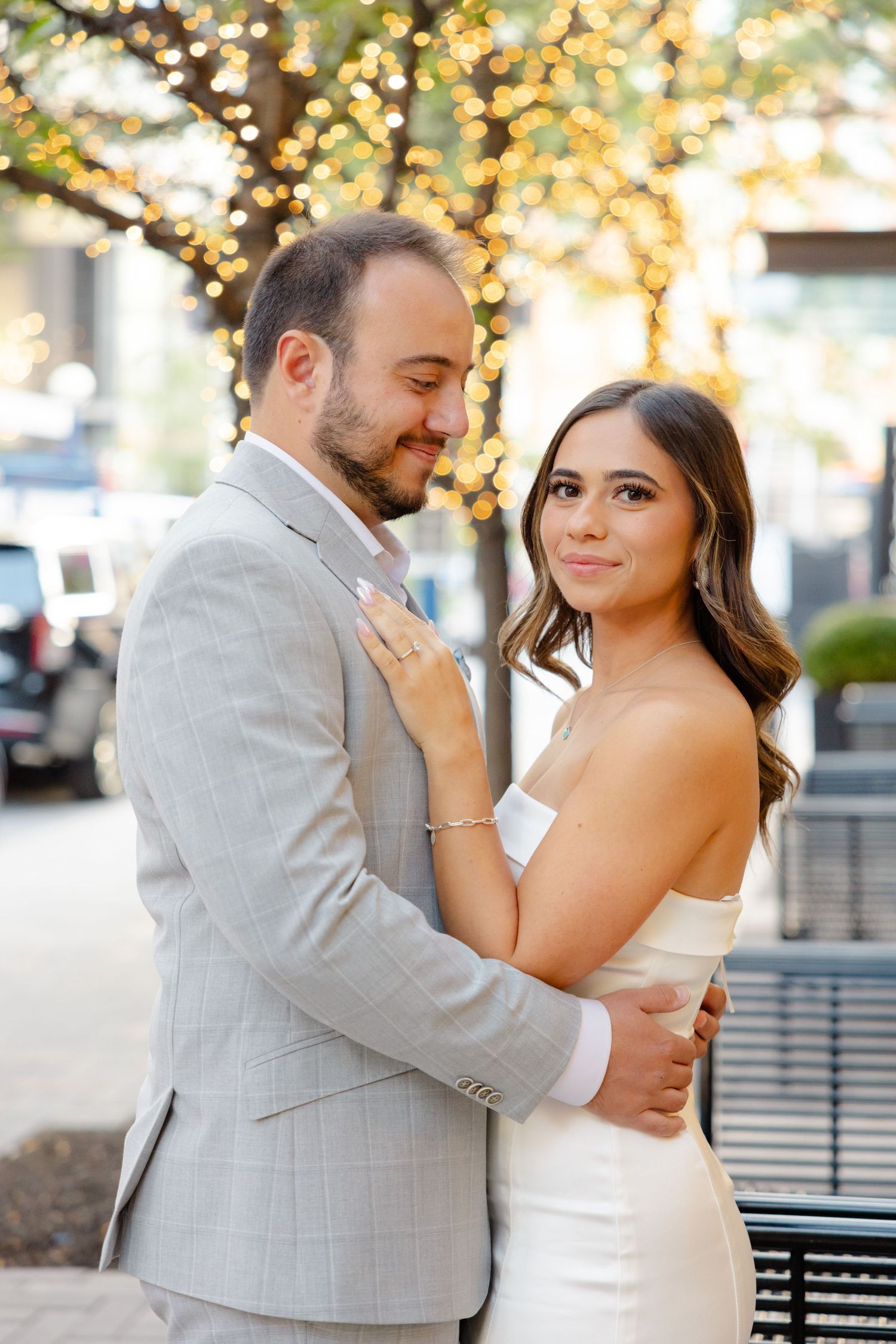 Romantic couple embraces outdoors with twinkling lights in background.