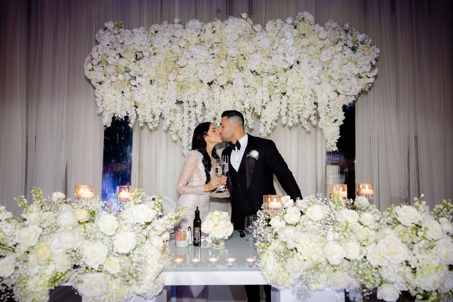 Couple shares champagne toast behind lavish white floral arrangements at wedding reception.