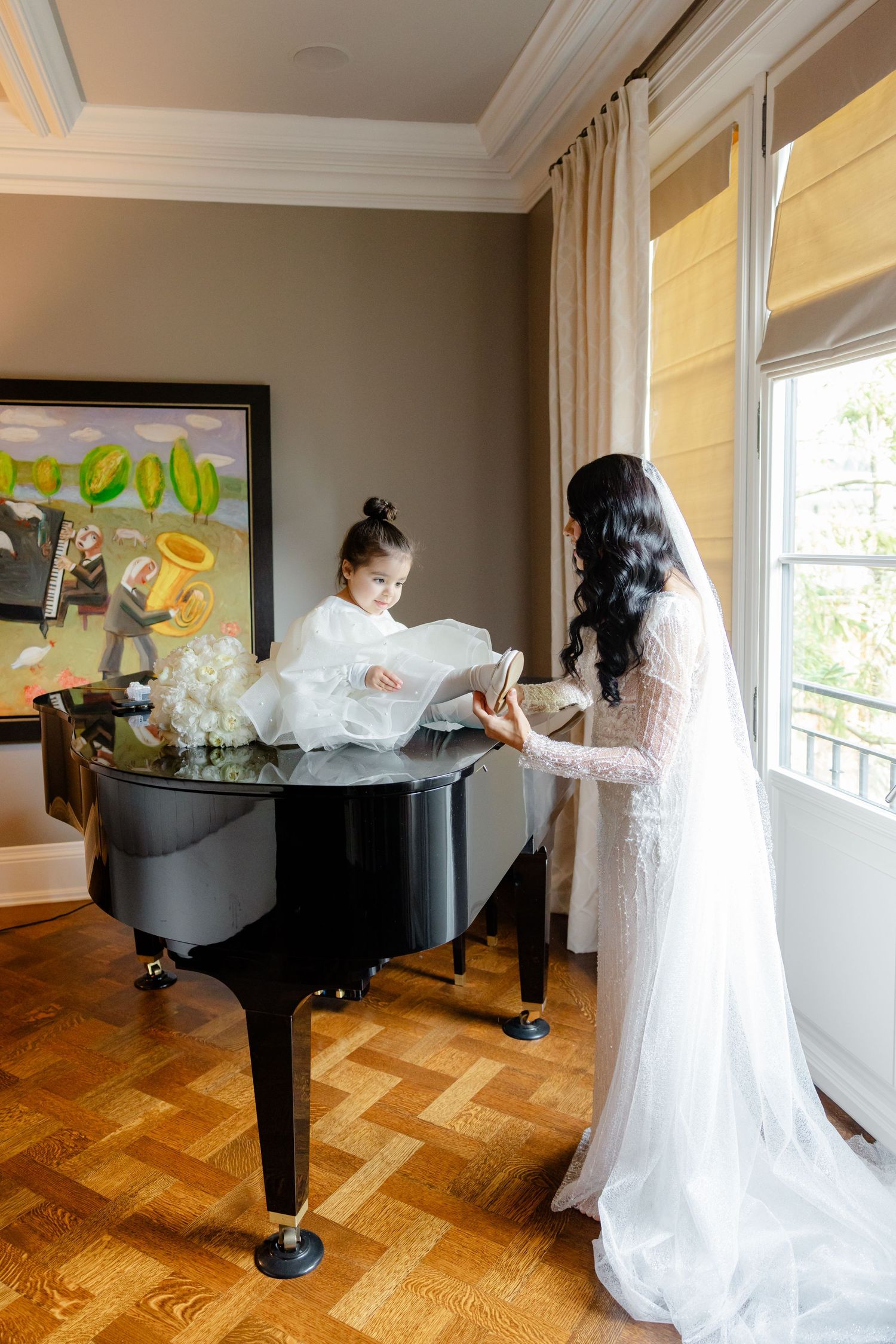 A bride in lace gown and small attendant in white dress stand by a piano near a window.