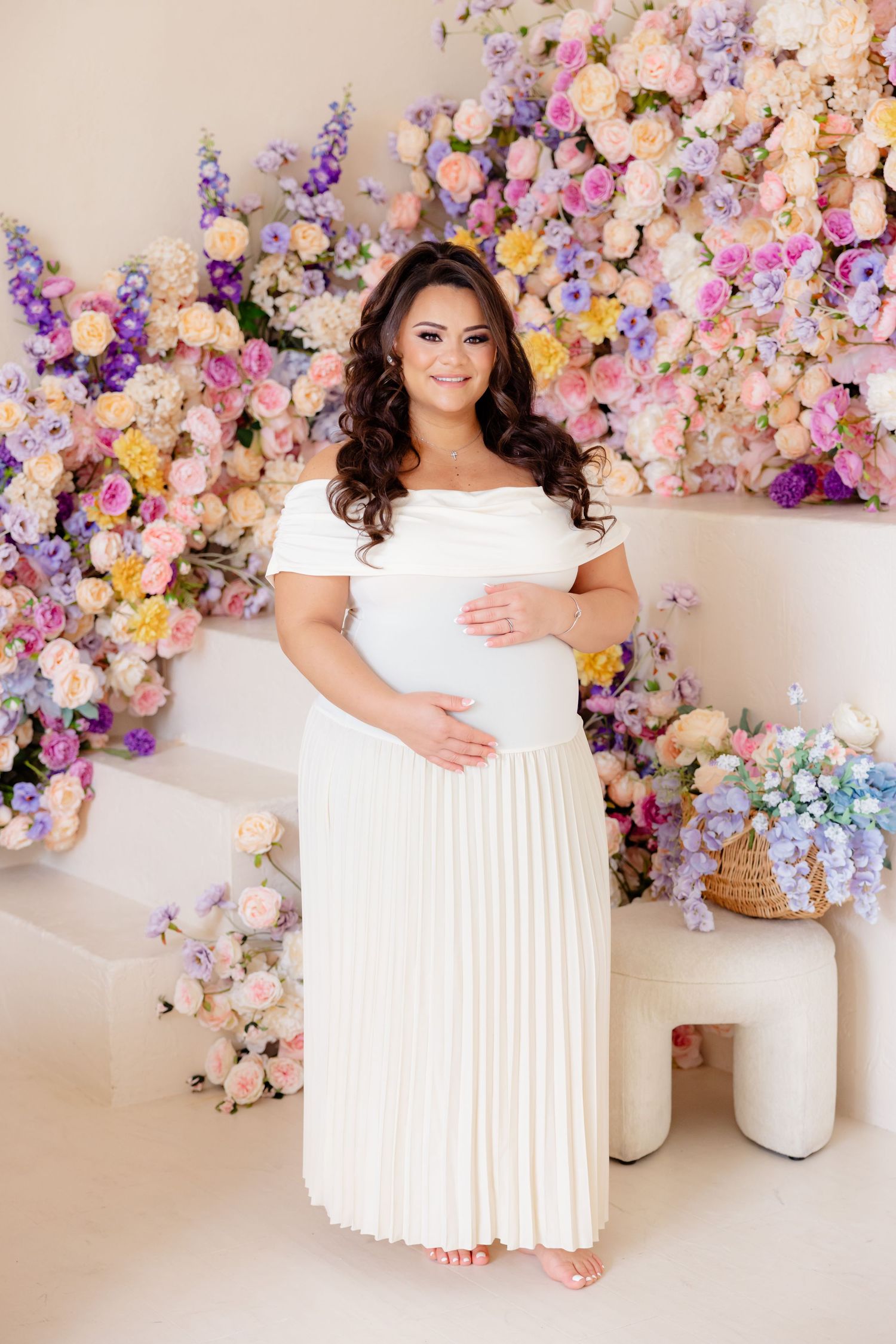 A pregnant woman in a white off-shoulder gown stands against a backdrop of pink, purple and peach floral arrangements.