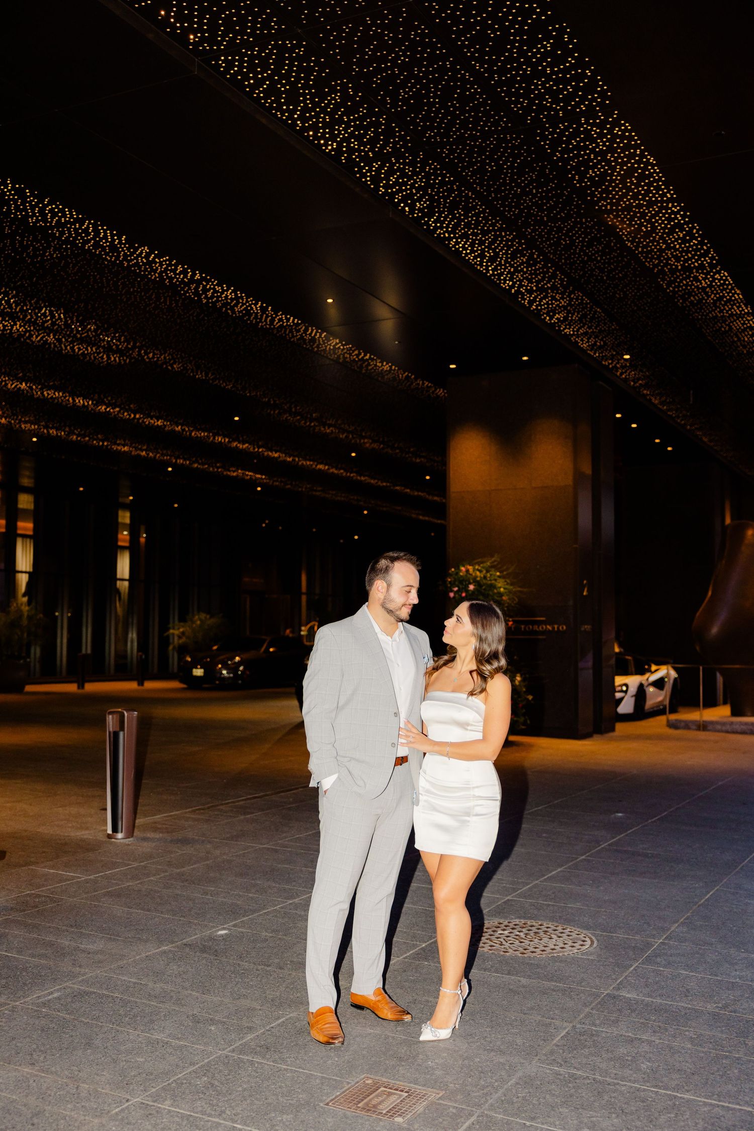 Elegant couple stands on outdoor plaza at night under decorative lighting.