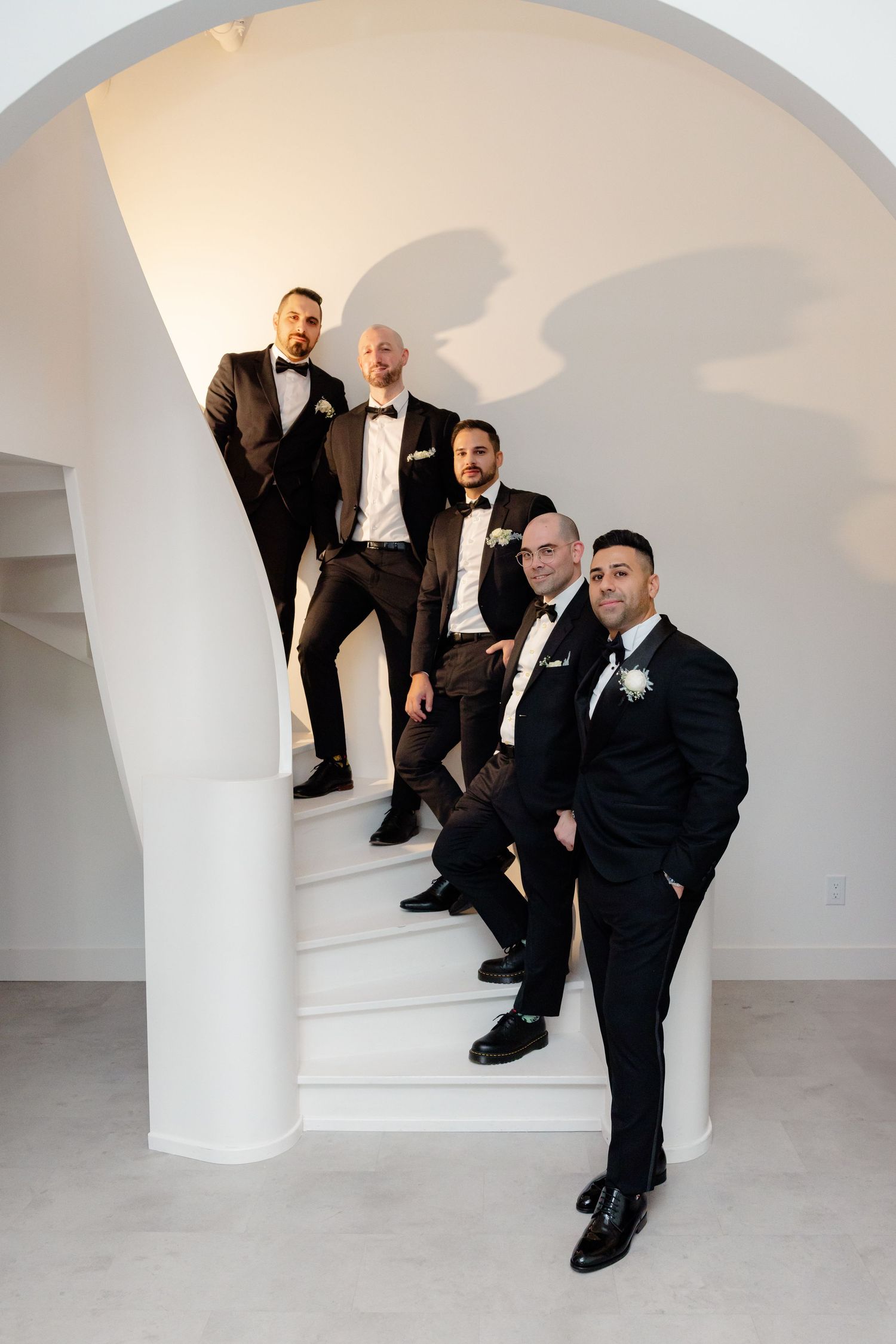 Groomsmen in black tuxedos posed on white spiral staircase in modern setting.