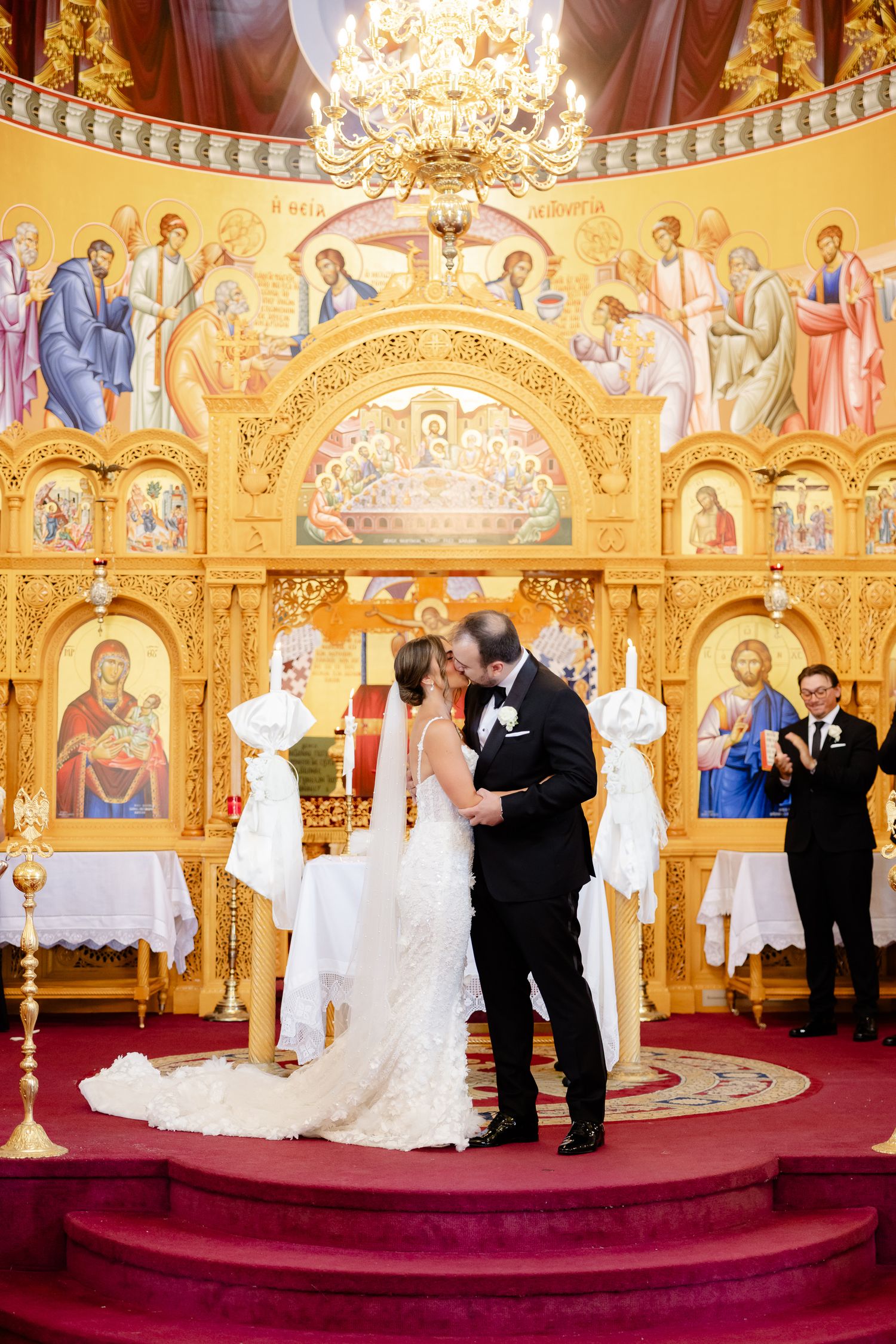 Ornate Orthodox church wedding scene with red carpeted steps, golden religious artwork, and chandeliers overhead.