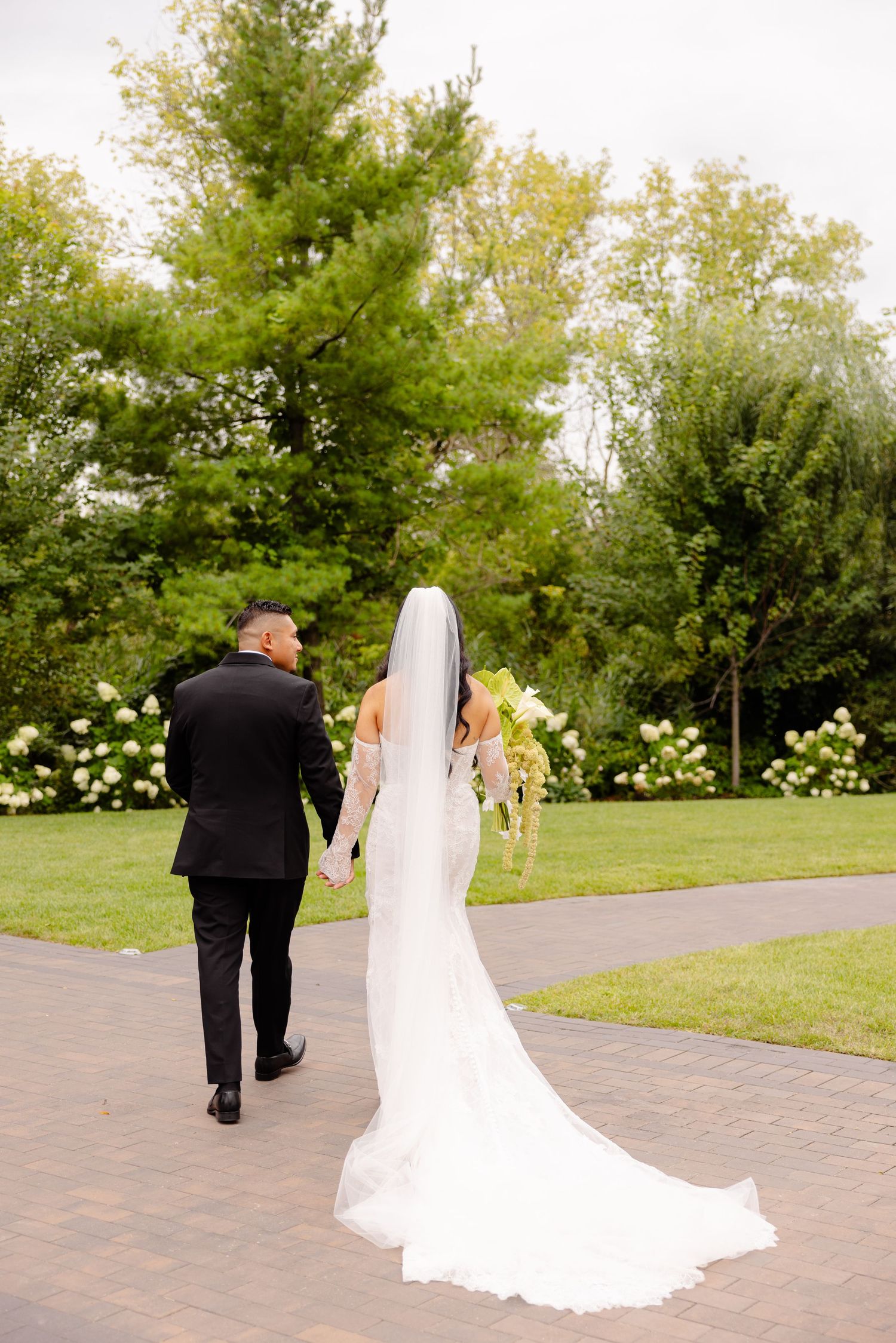 Wedding couple walking along a brick pathway surrounded by lush green trees and landscaping on a beautiful summer day.