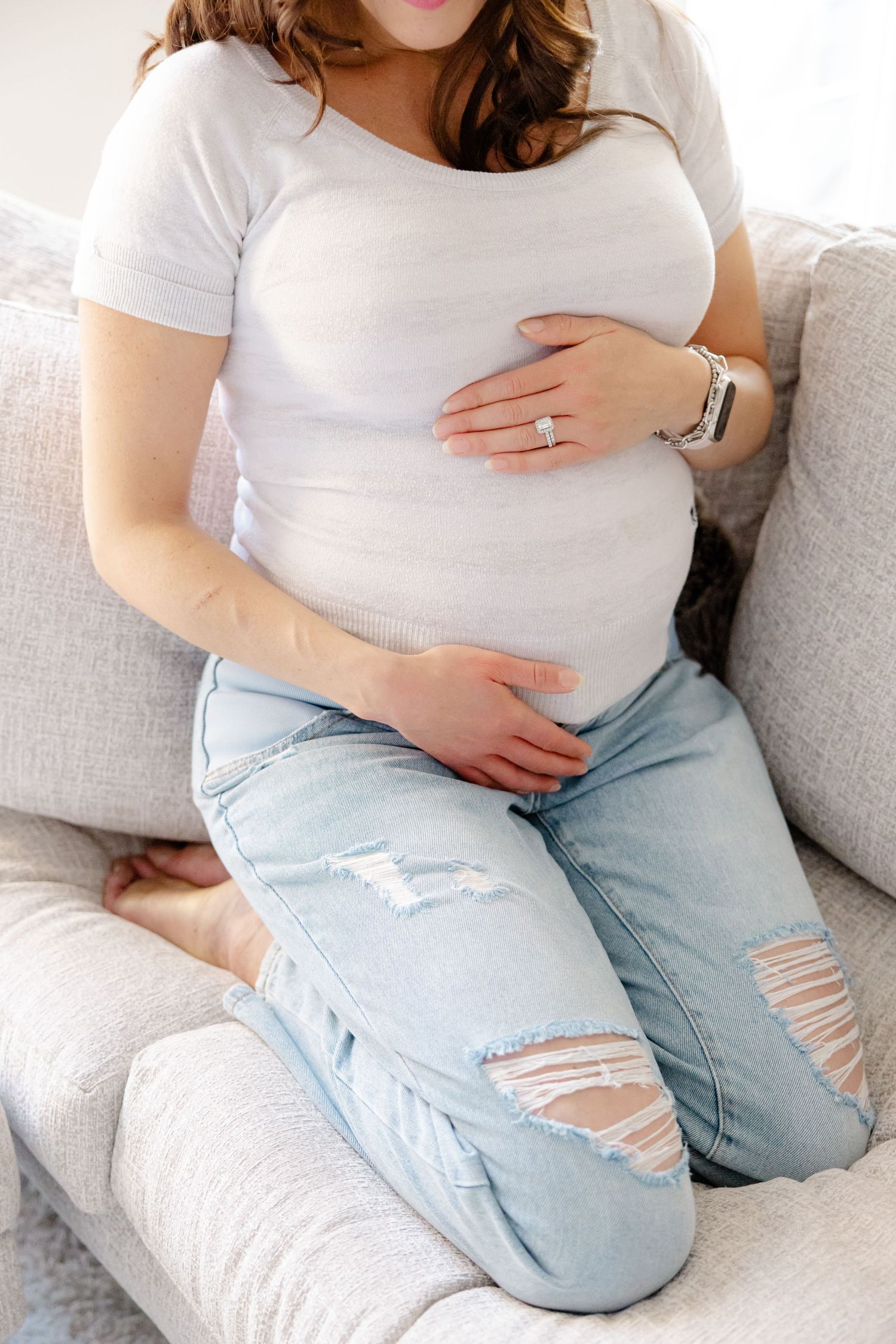A pregnant woman in a white shirt and ripped jeans sits on a gray couch.