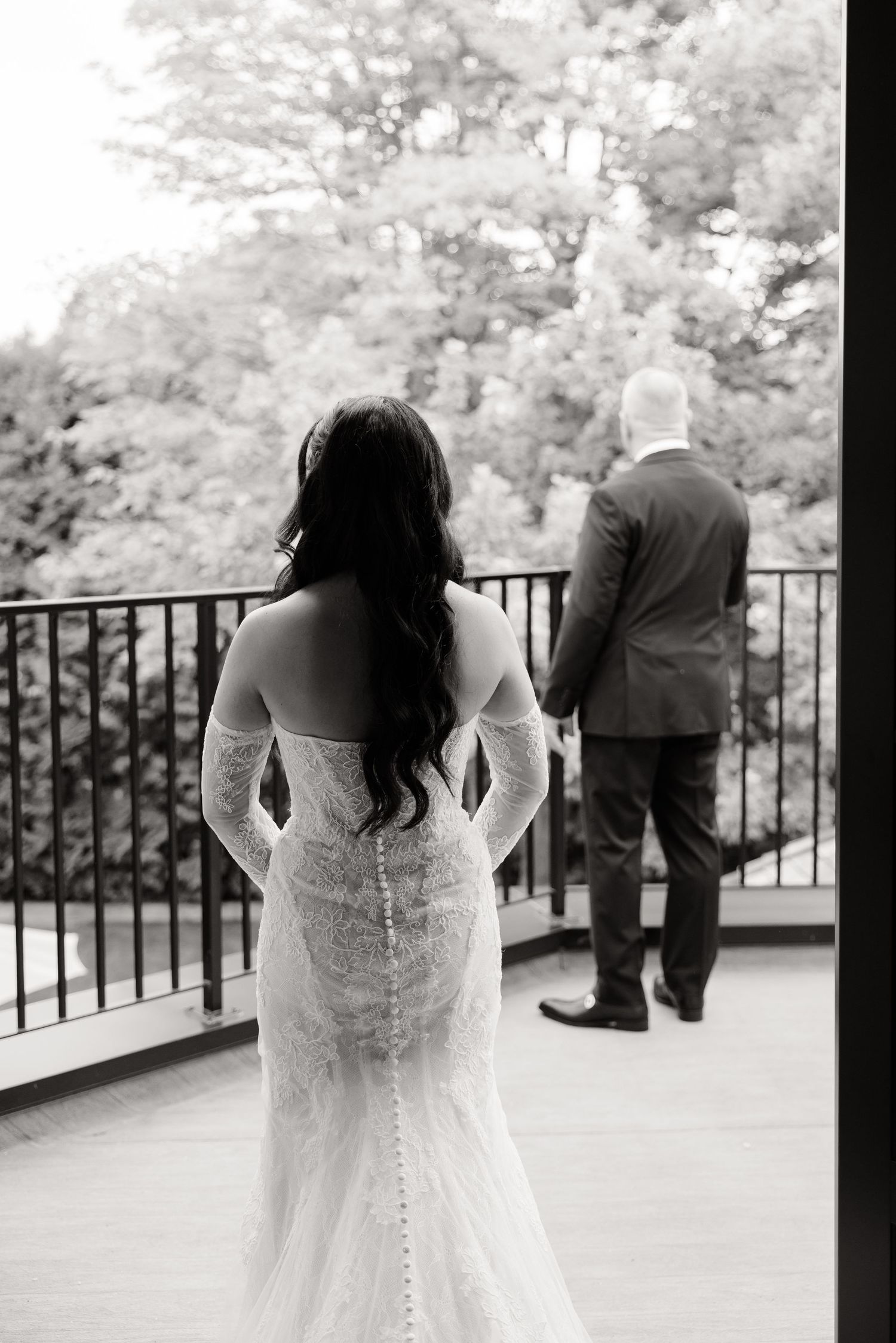 A bride in a white lace wedding dress stands on a balcony while her groom looks out over the railing in the distance.