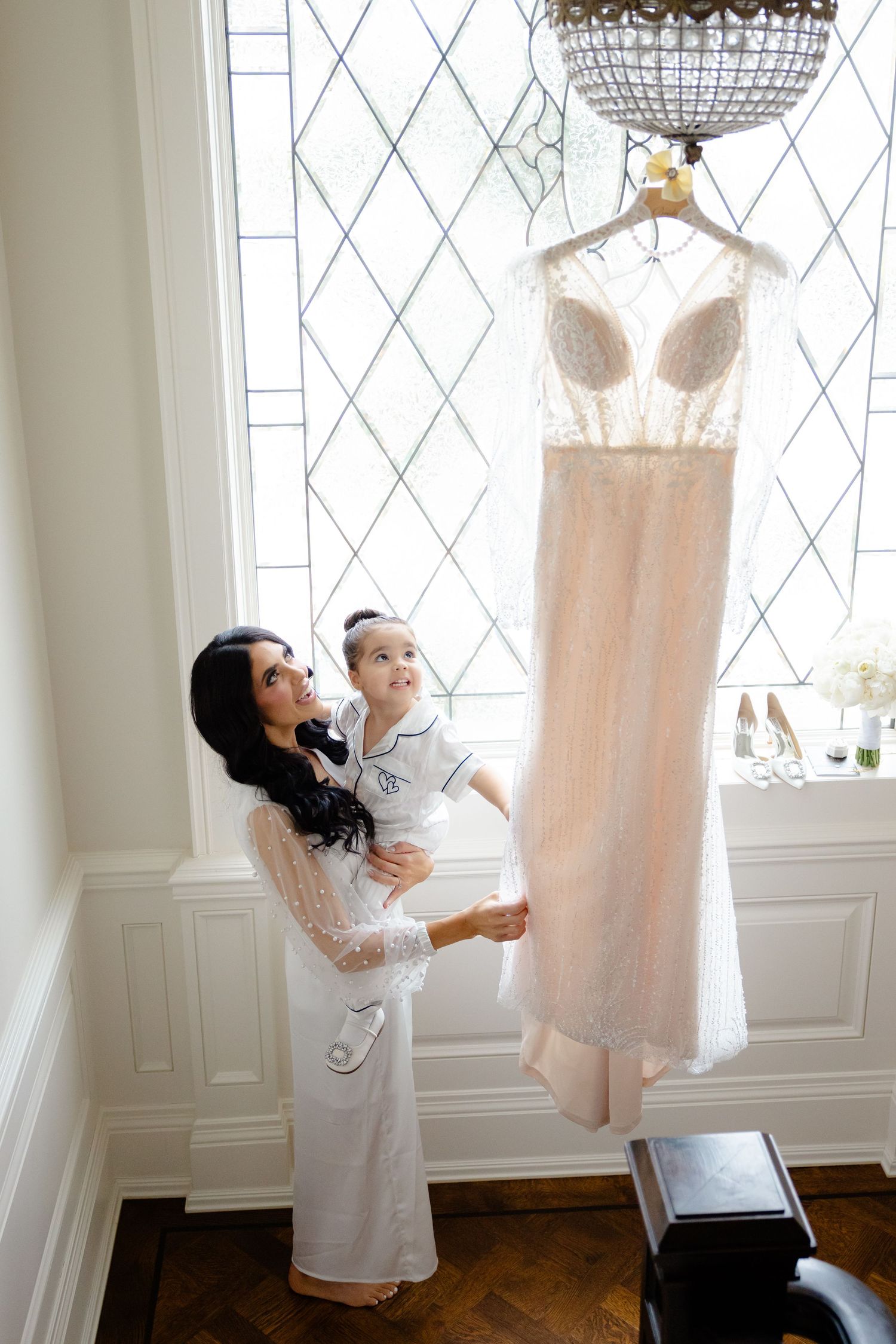 Wedding dress hangs elegantly from chandelier as two people share a tender moment below.