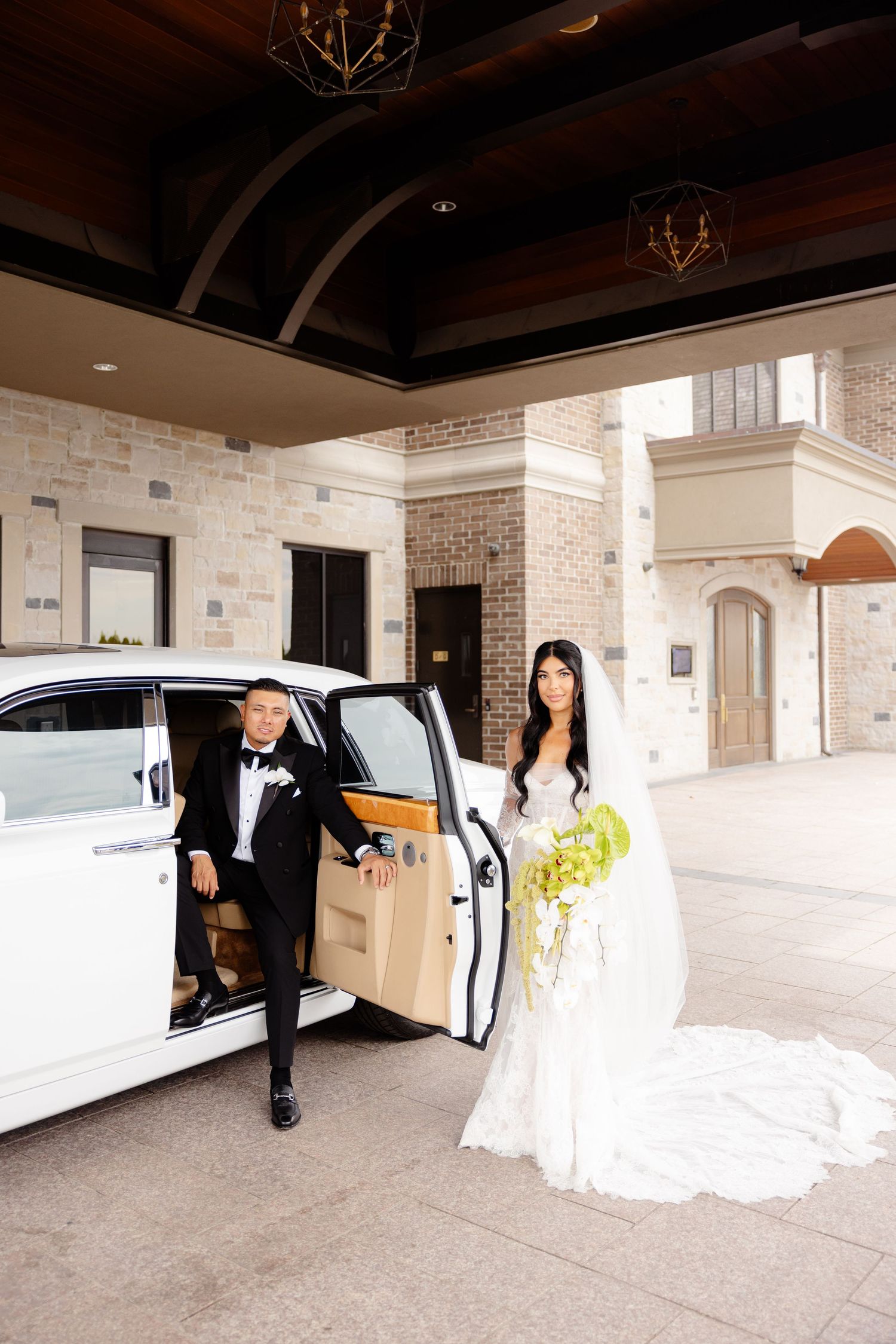 A couple arrives at an elegant stone building entrance in formal attire with a white stretch limousine.