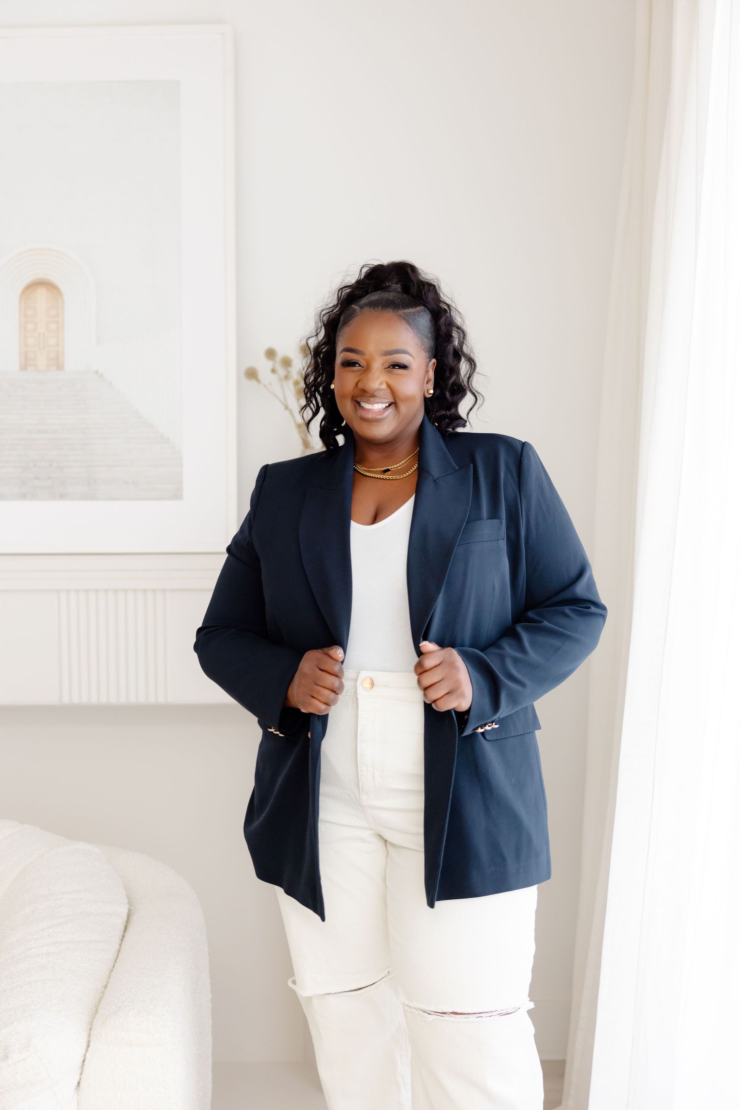 Professional portrait of someone wearing a navy blazer with white pants and top in a bright minimalist interior setting.