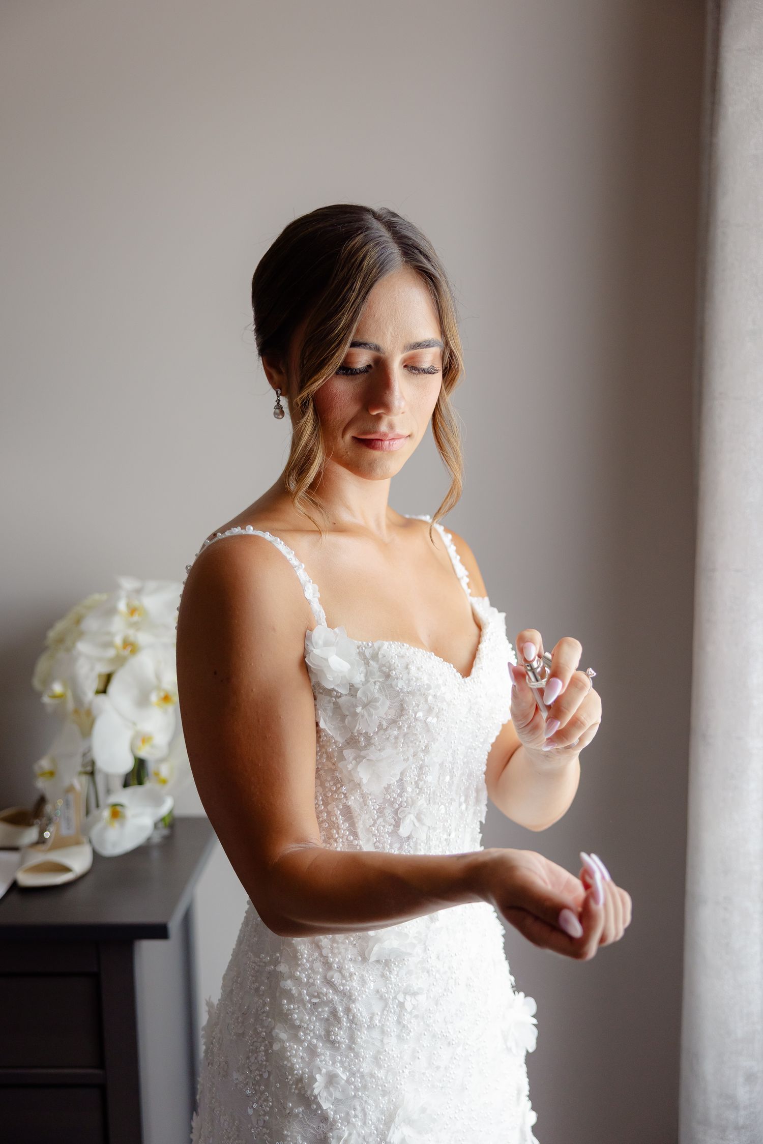 A person in a white lace wedding dress sprays perfume in a softly lit room with orchids visible in the background.