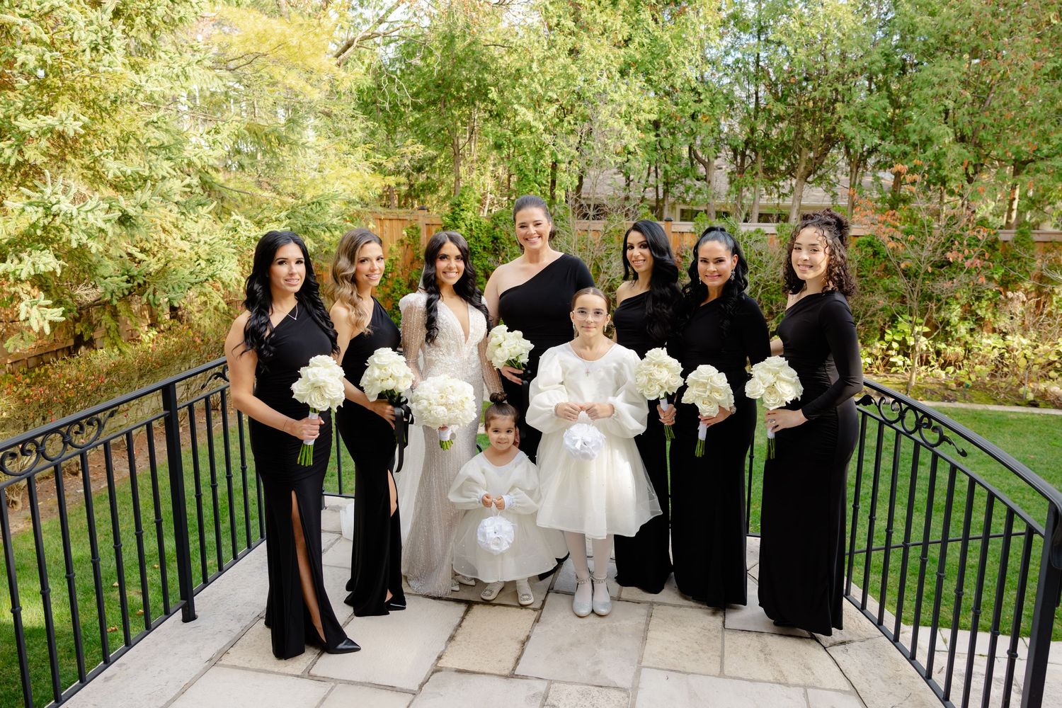Wedding party in black dresses poses with bride and flower girl on outdoor stone terrace.