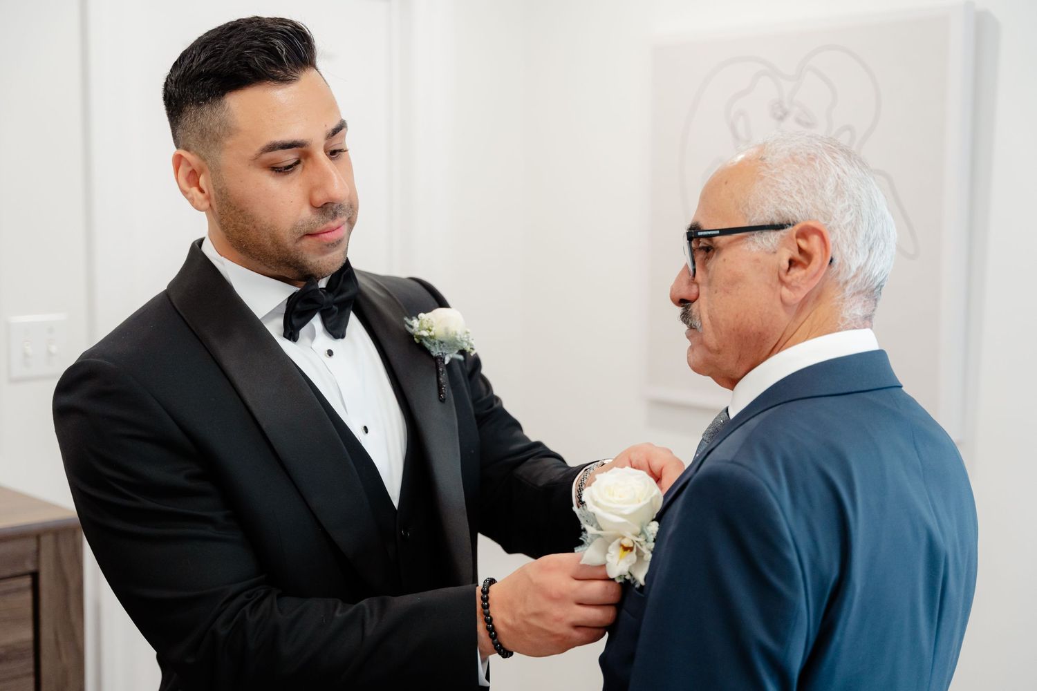 A touching pre-wedding moment as two men help each other with white boutonnieres.