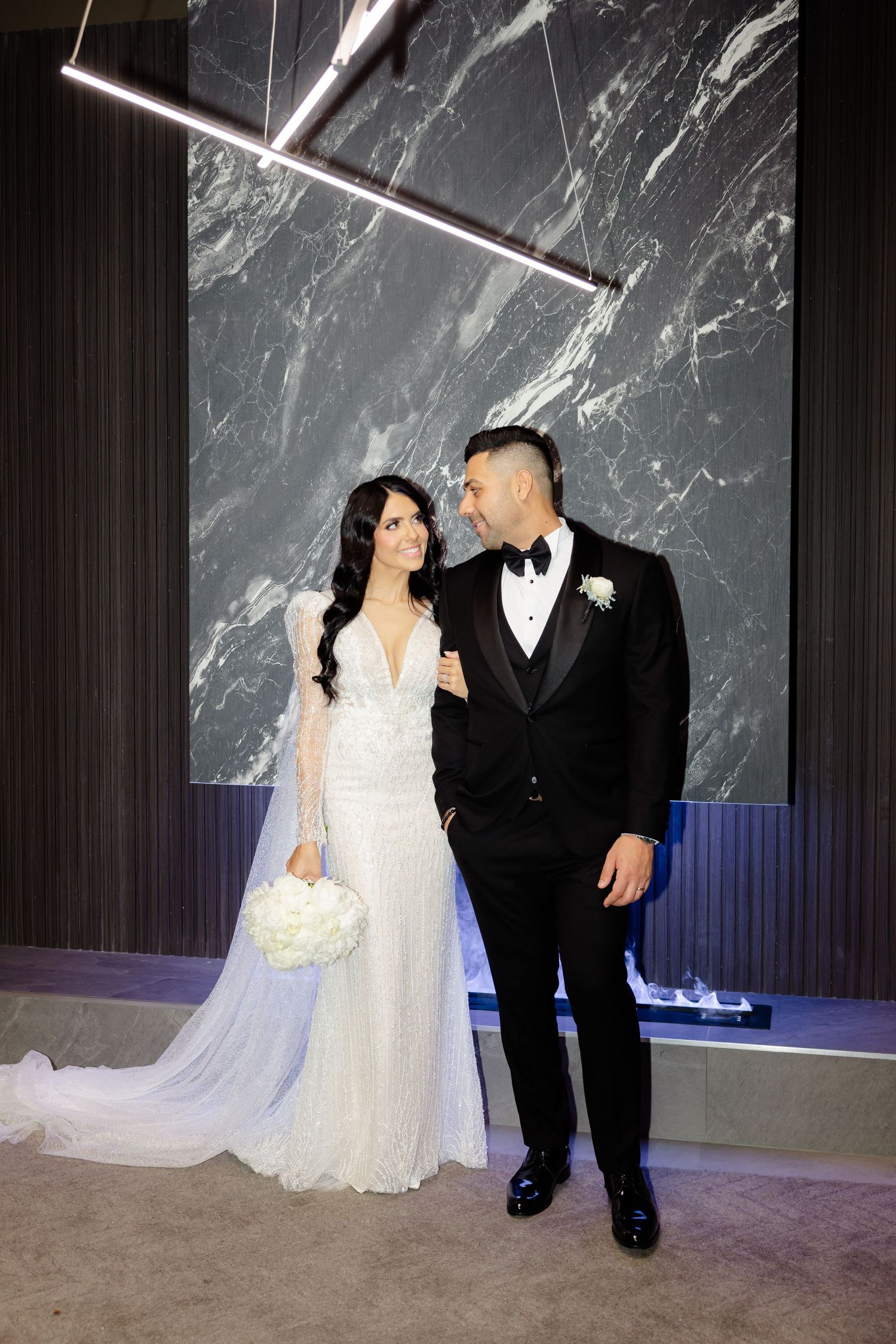 Bride in lace gown and groom in tuxedo pose against marble backdrop with modern lighting.