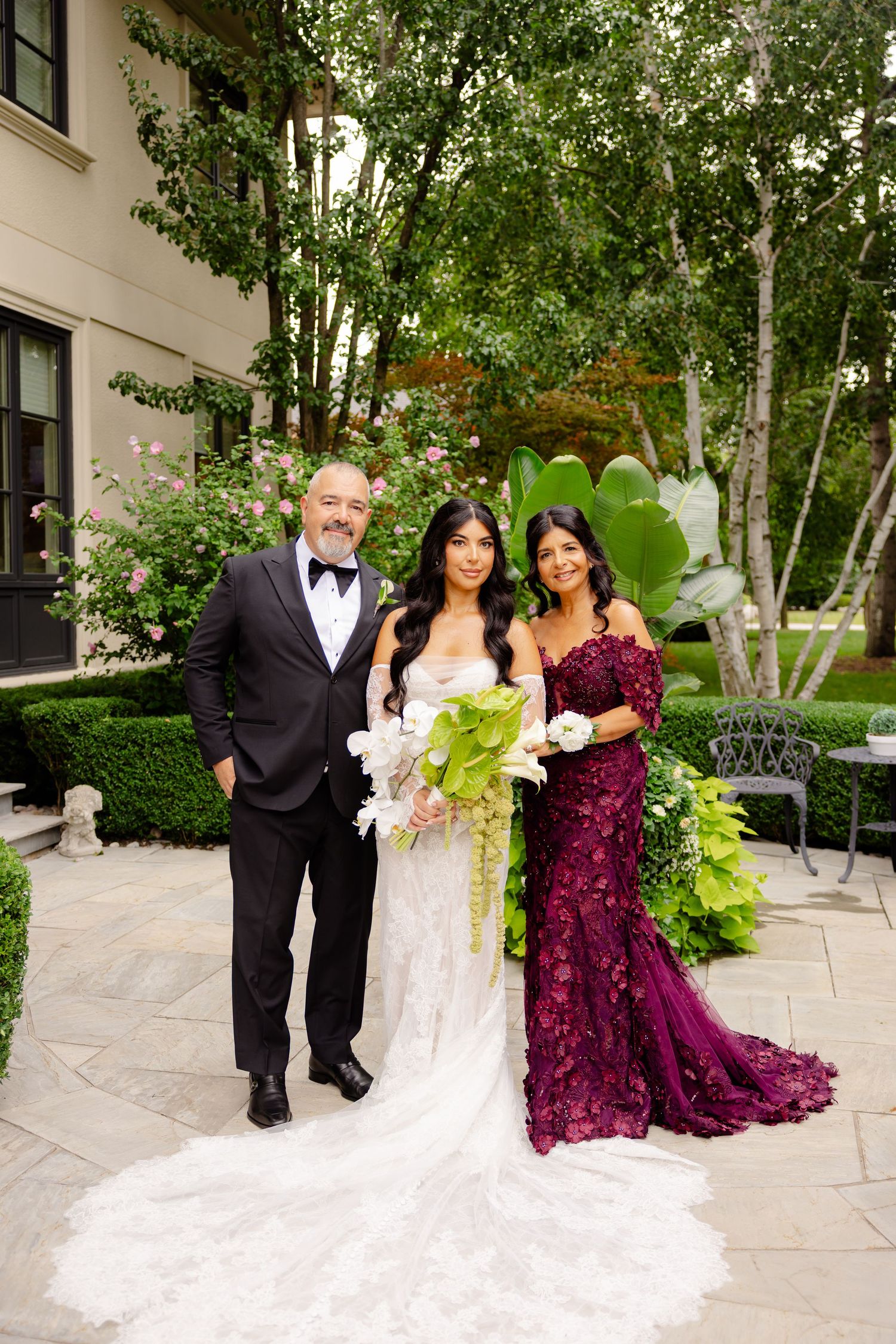 Wedding party poses in formal attire against garden backdrop with birch trees and manicured landscaping.