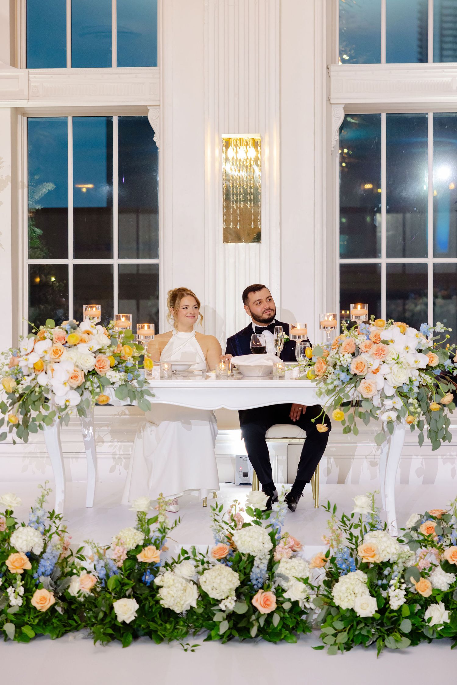 Elegant wedding reception setup with lush floral arrangements in peach and white along tables in a bright white room.