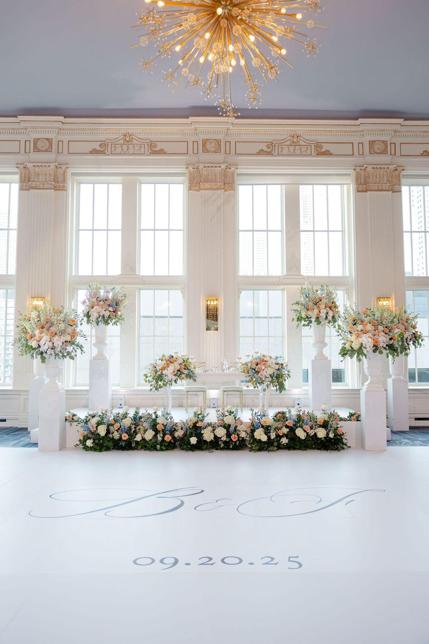 Grand ballroom with starburst chandelier and floral arrangements along windows for wedding reception.