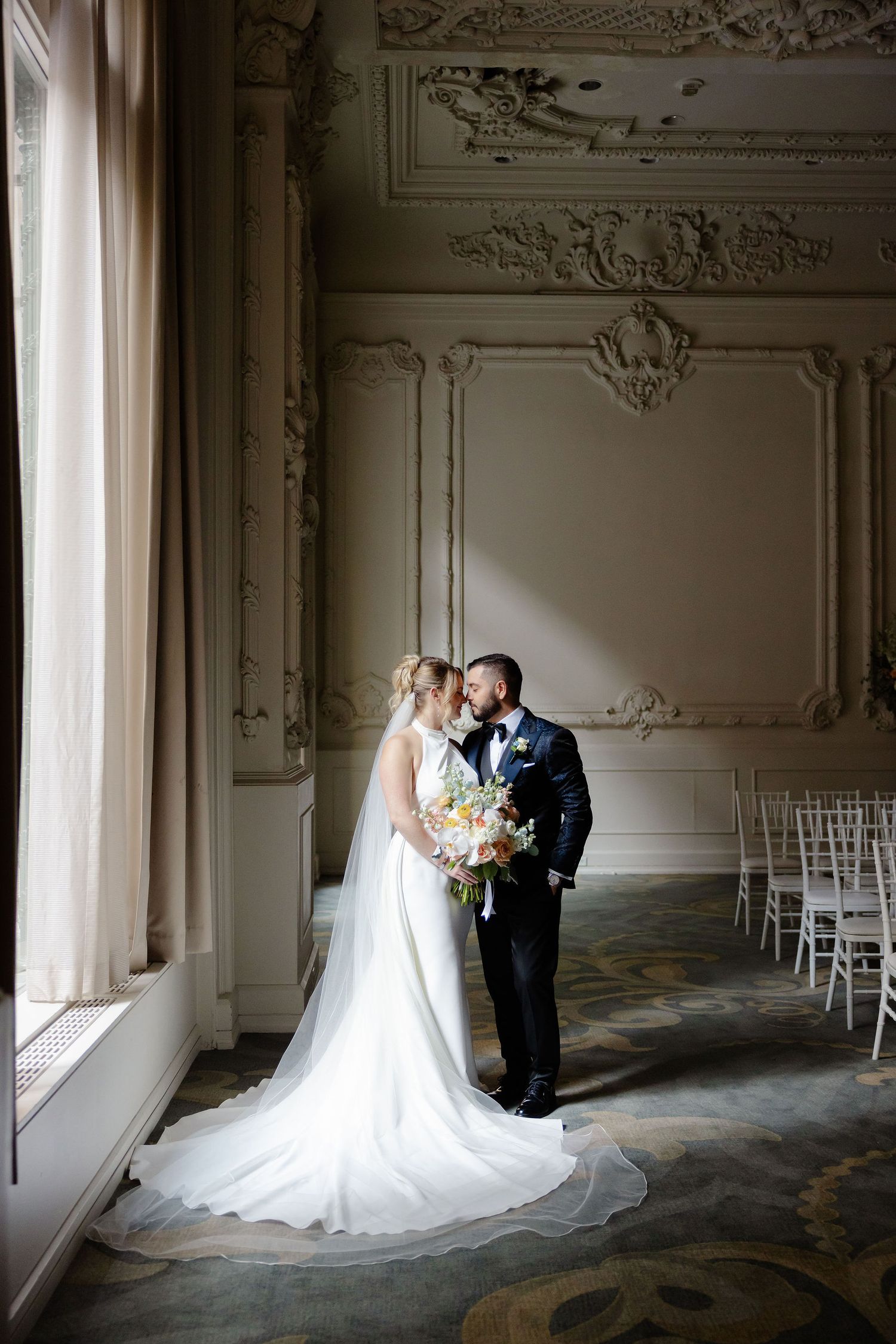 Wedding couple poses in a grand ballroom with ornate architectural details and dramatic lighting.