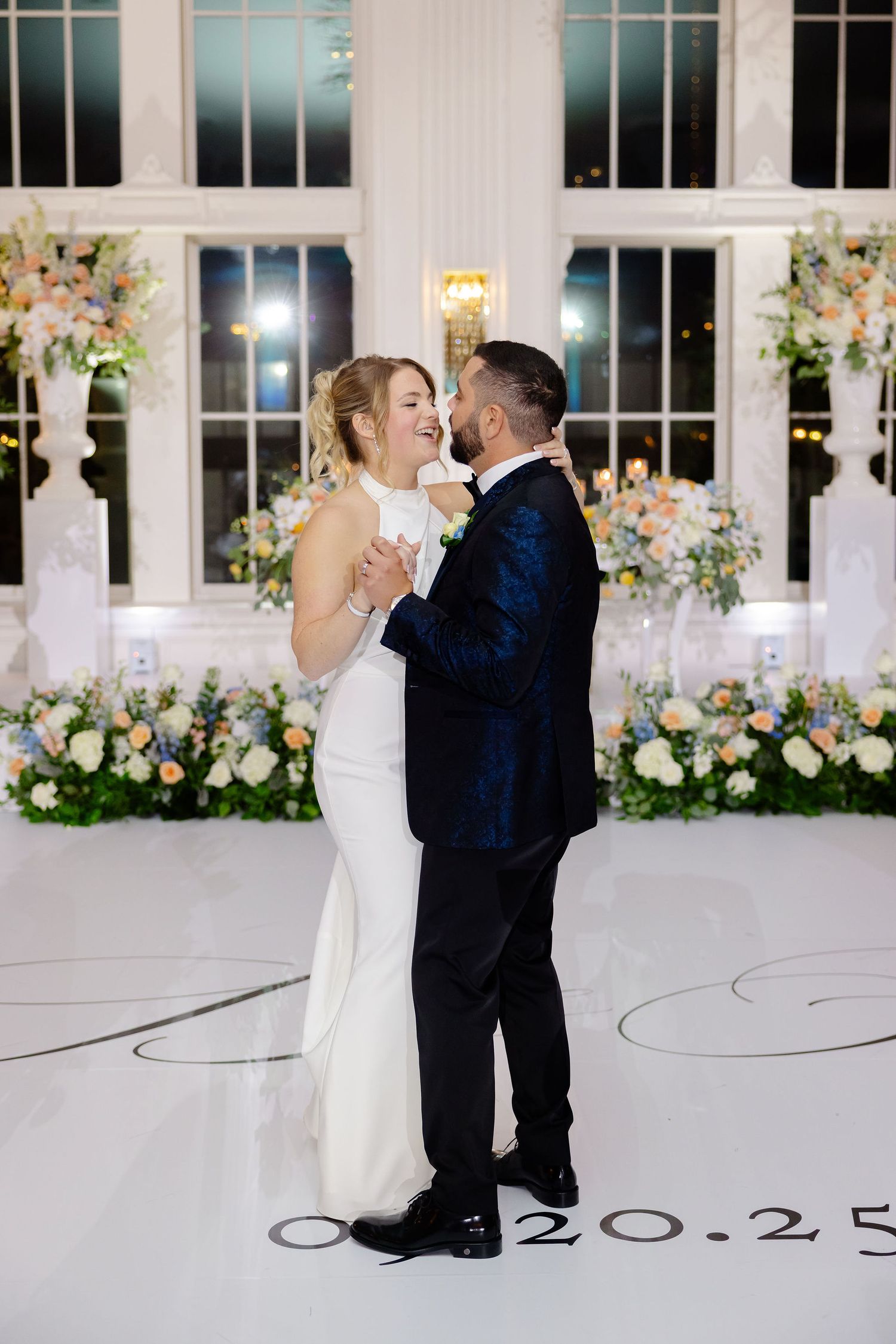 Newlyweds share an intimate moment on their wedding dance floor surrounded by elegant floral arrangements and tall windows.