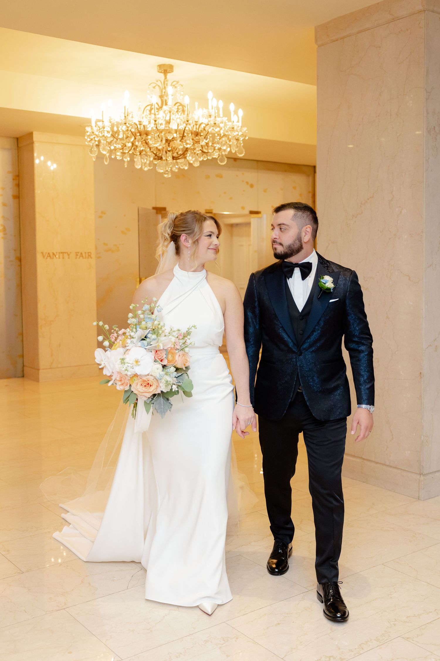 Newlyweds walk together through an elegant hallway with crystal chandeliers overhead.