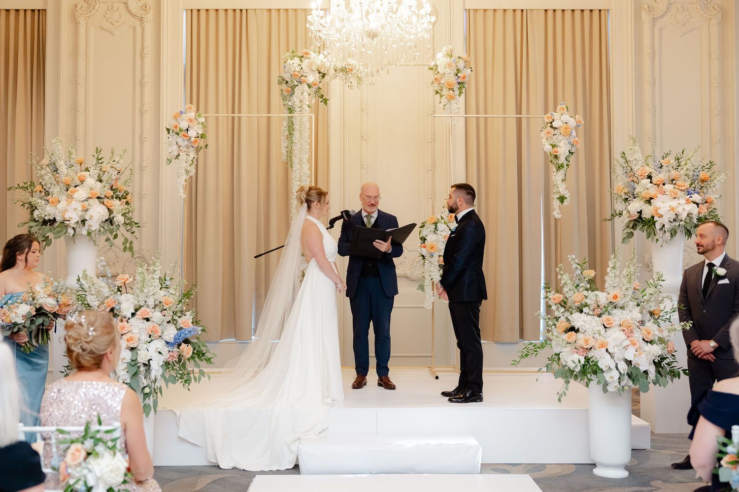 Luxurious wedding ceremony setup with crystal chandelier and floral arrangements in an ornate ballroom.