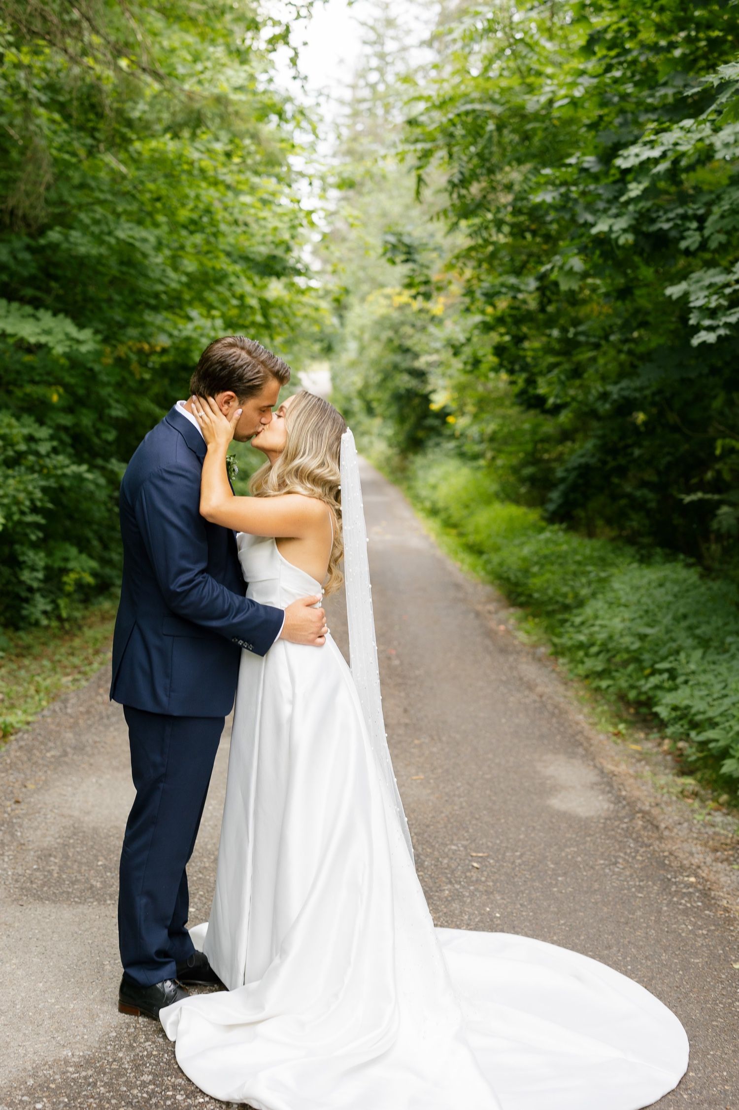 Intimate wedding moment captured on tree-lined path with flowing white dress train.