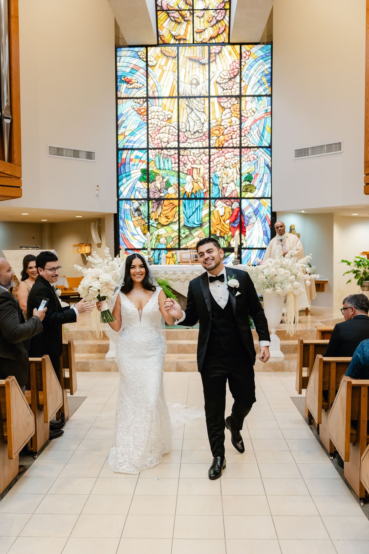 Joyful couple walking down church aisle after ceremony with stained glass window behind.