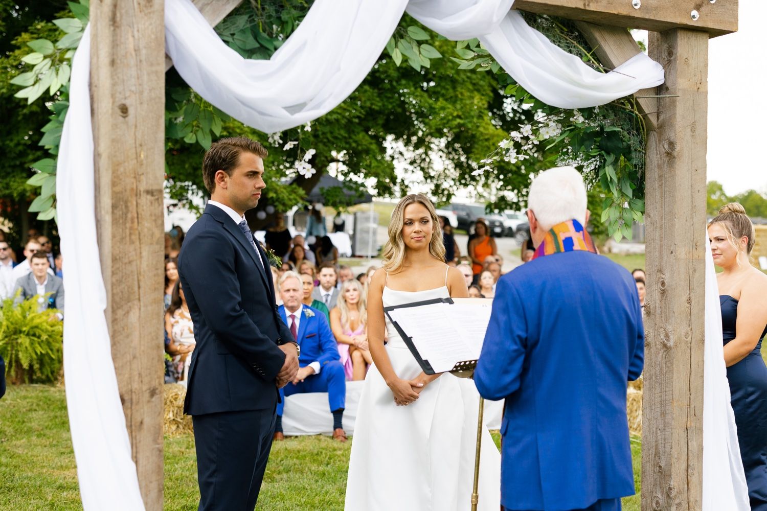 An outdoor wedding ceremony takes place under white fabric draped wooden posts.