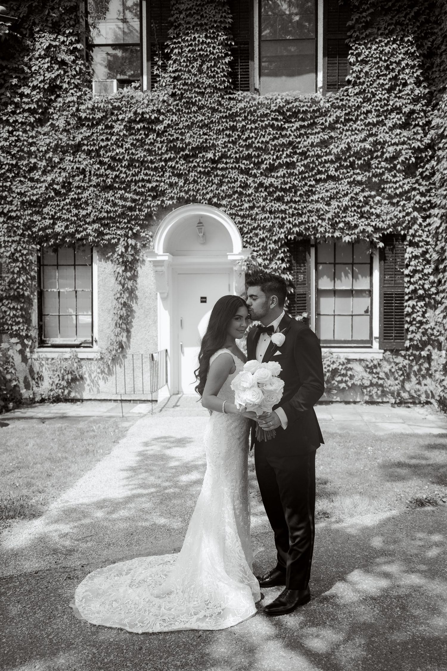 Black and white photo of couple kissing in front of ivy-covered building with arched entrance.